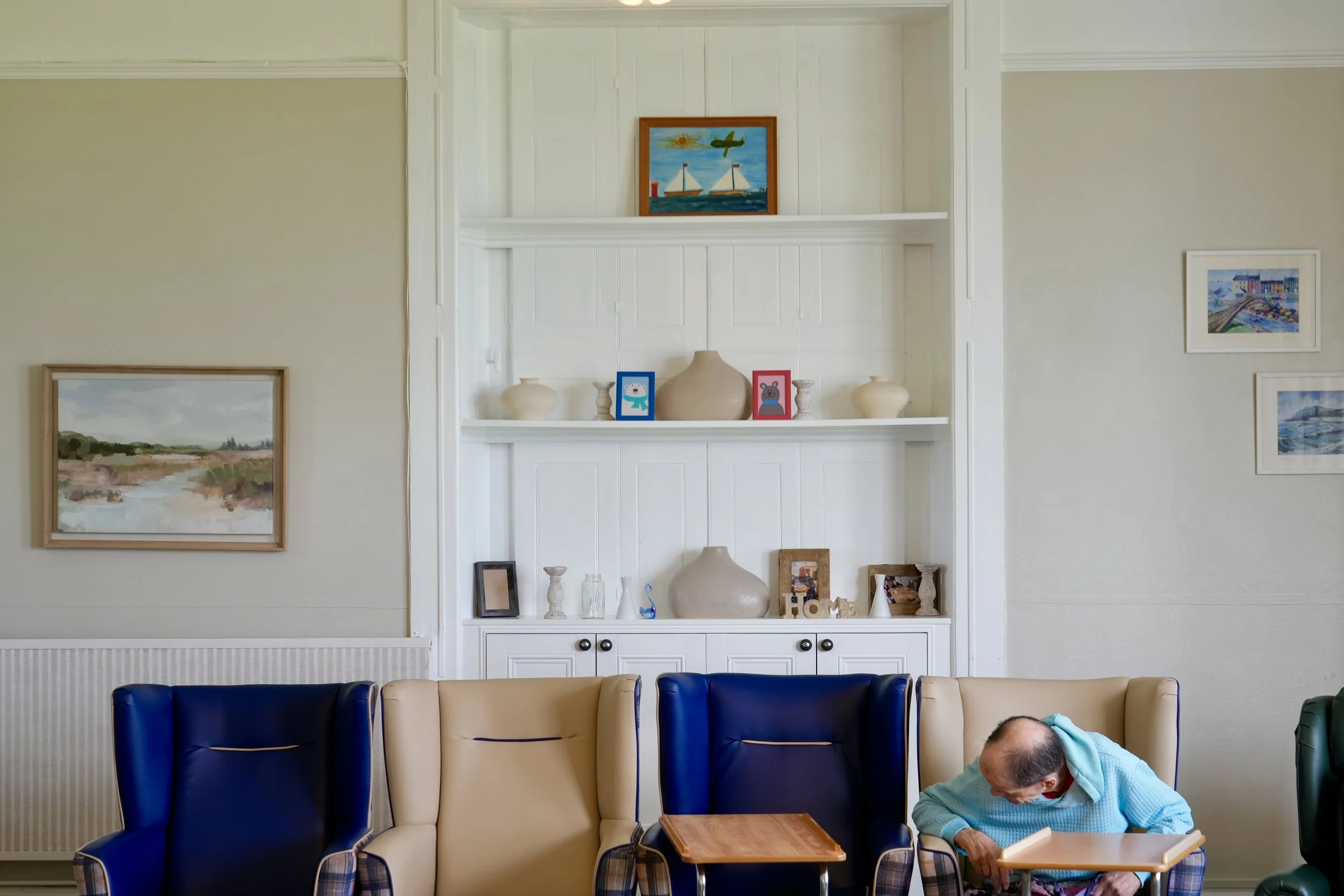 A man with a shaved head and wearing a light blue hoodie sitting in a blue and beige armchair, leaning over a small wooden table in a room with framed artwork on the cream-colored wall and blue and tan chairs in front.