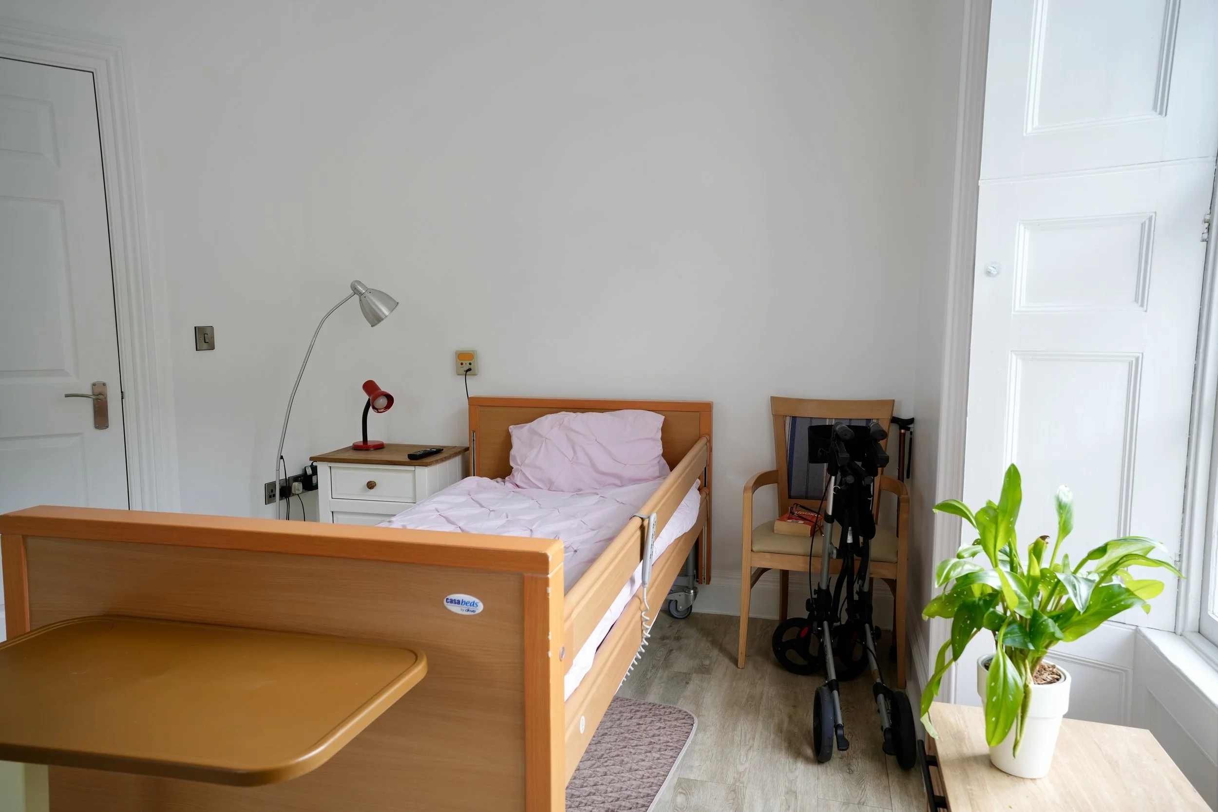 A bedroom with a wooden bed and pink bedding, a white bedside table with a red lamp, a gray wall lamp, a treadmill, a chair, a telescope, and a potted plant near a window.