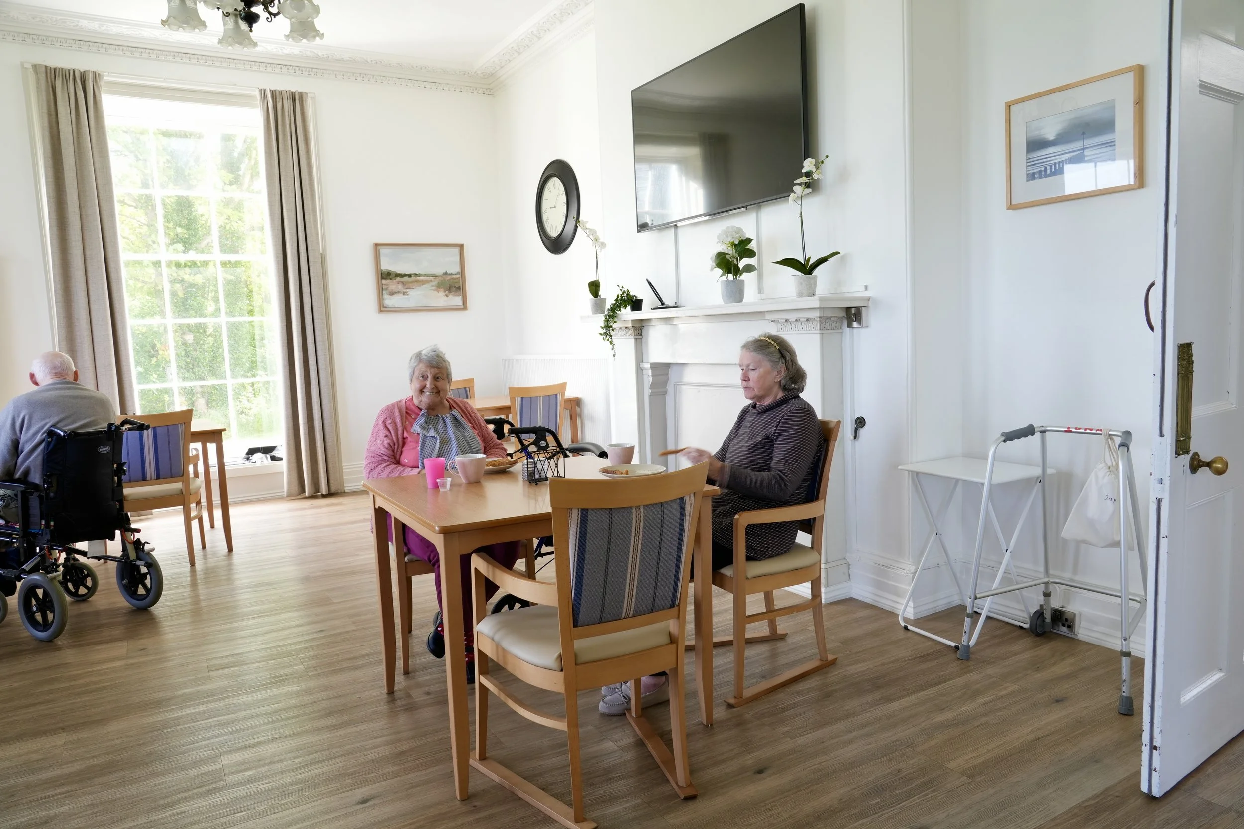 Senior women sitting at a dining table in a bright room, with large window behind, decorated with framed artwork and plants, and a television mounted on the wall.