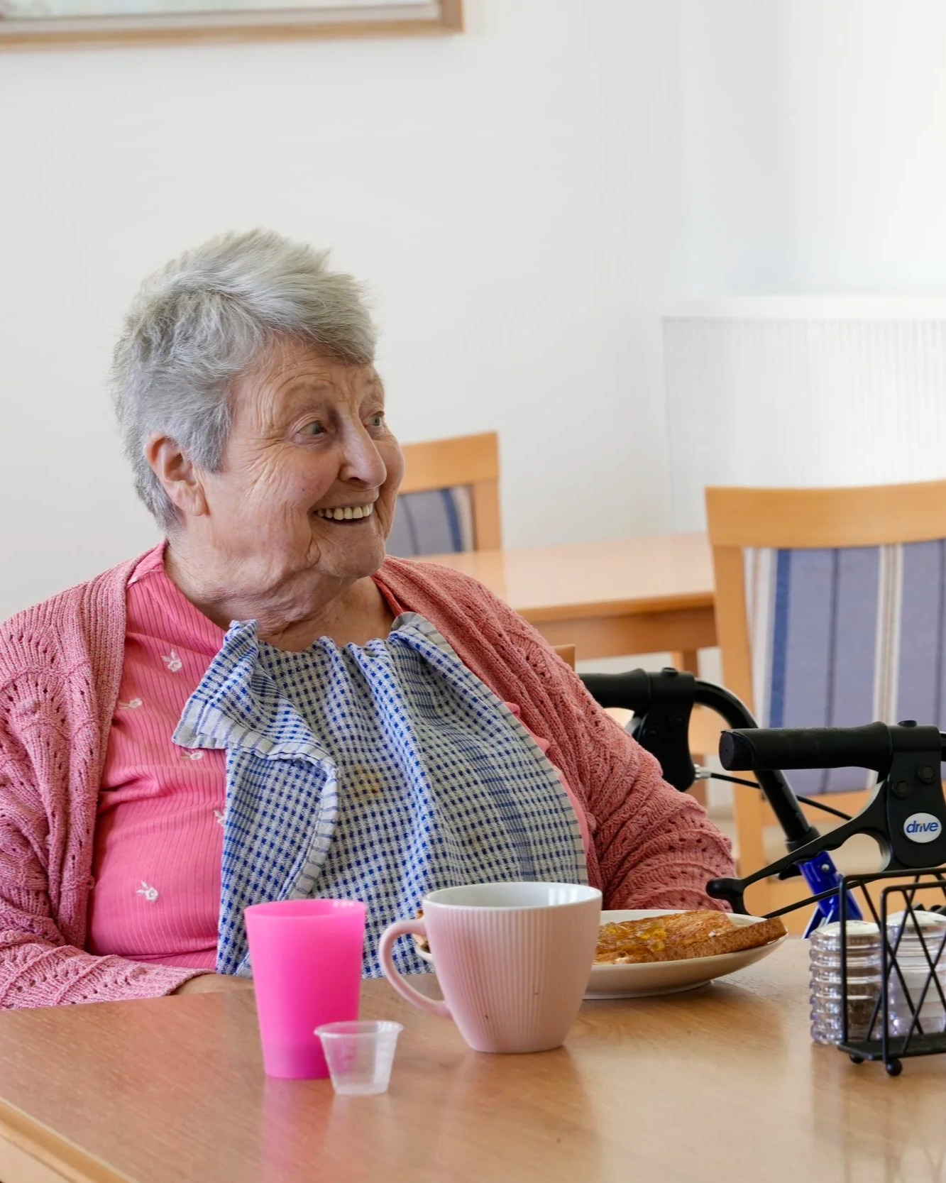 An elderly woman with gray hair smiling, seated at a table with food and drinks, wearing a pink shirt, a patterned bib, and a pink cardigan, in a dining room.
