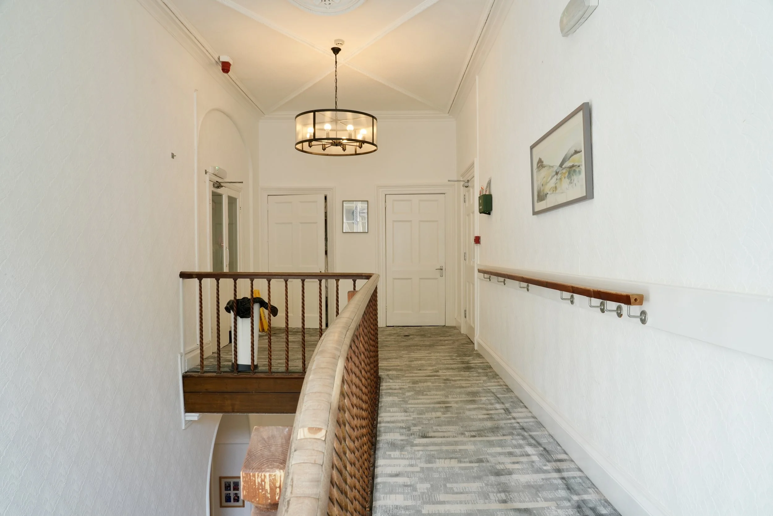 An indoor hallway with white walls, a wooden handrail on the right side, a modern circular chandelier hanging from the ceiling, multiple white doors at the end of the hall, framed artwork on the right wall, a green box on the wall, and a security cam