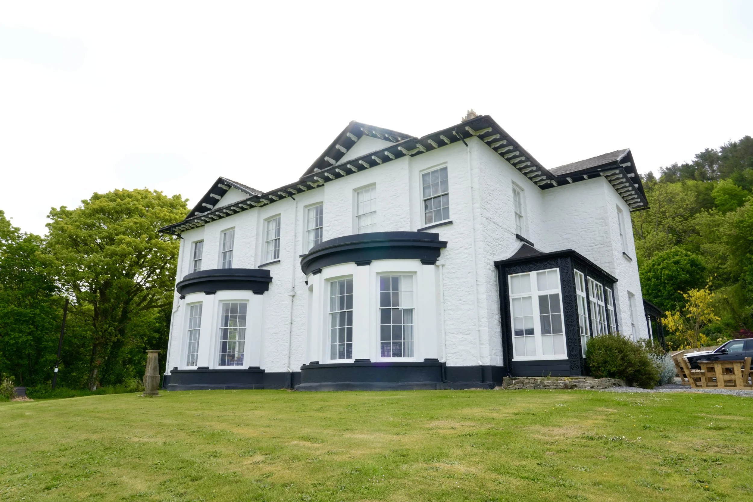 A large white house with black trim and multiple stories is surrounded by green trees and a well-maintained lawn.