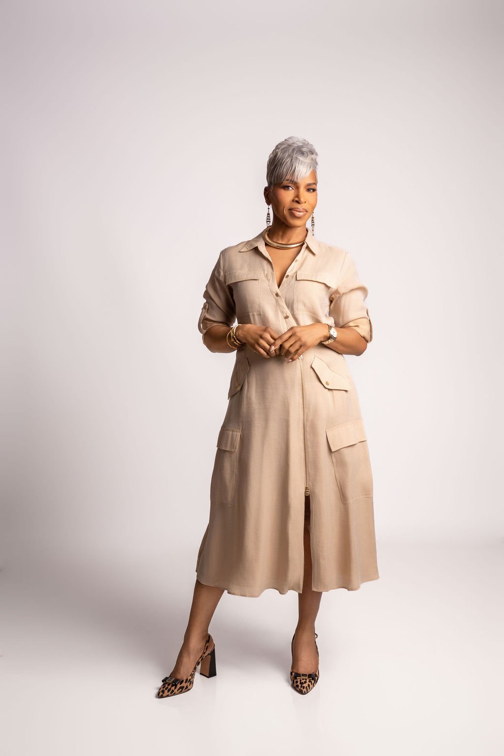 Stylish woman with short gray hair wearing beige dress, leopard print high heels, jewelry, and standing confidently against a plain white background.