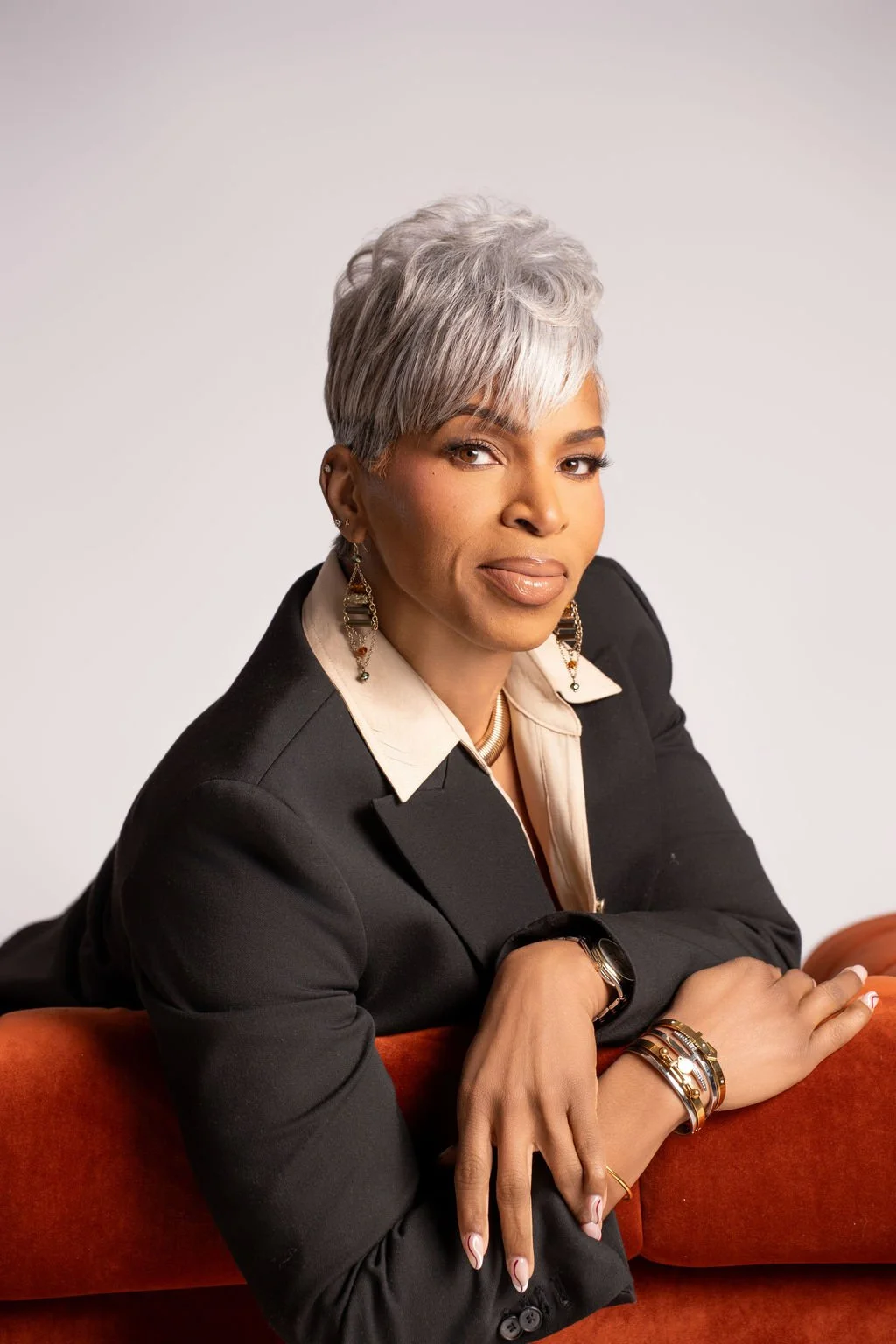 A professional woman with short gray hair wearing a black blazer and jewelry, sitting on an orange couch against a white background.