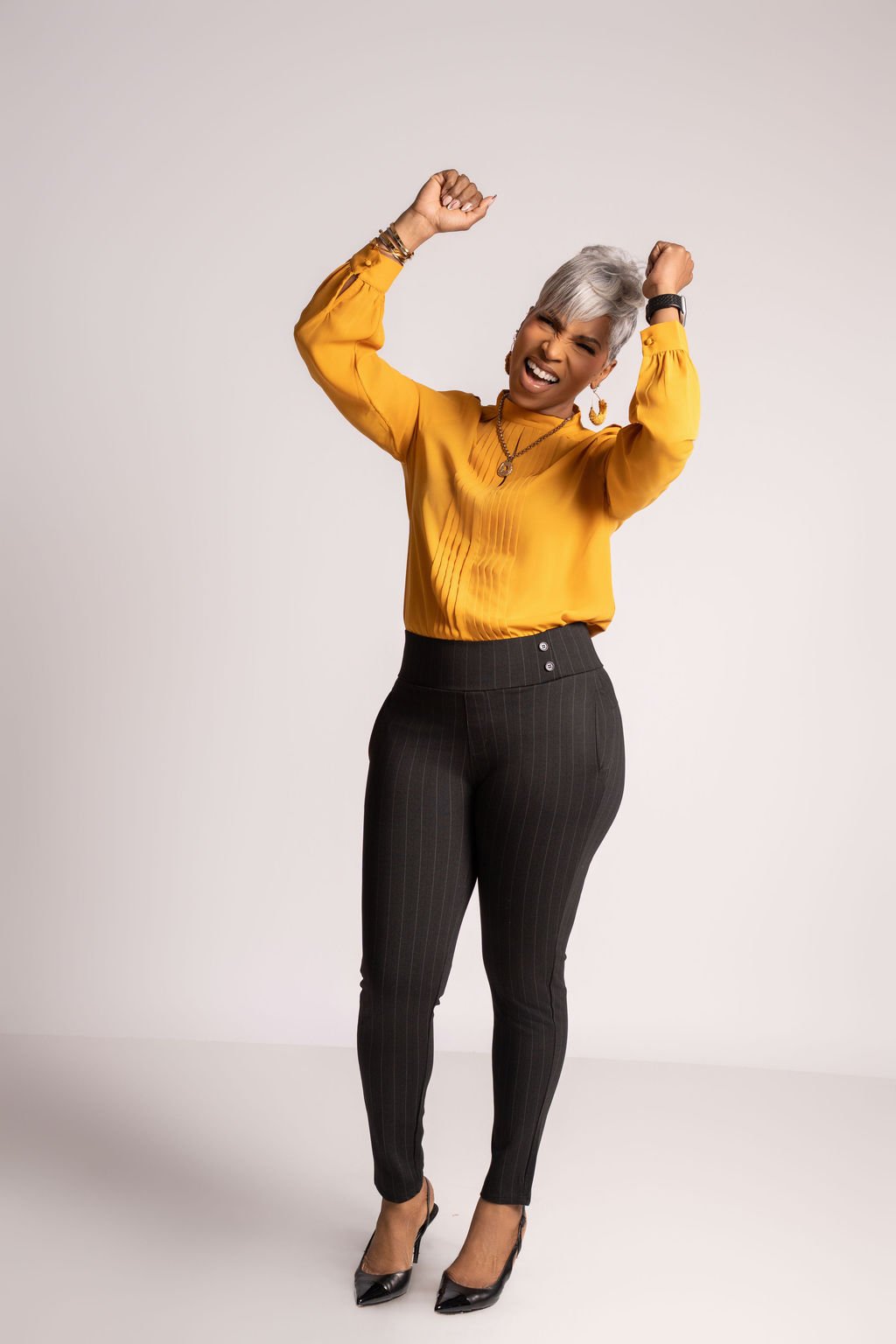 A woman with short gray hair wearing a yellow blouse and black pinstripe pants, standing against a plain background, smiling and showing excitement.