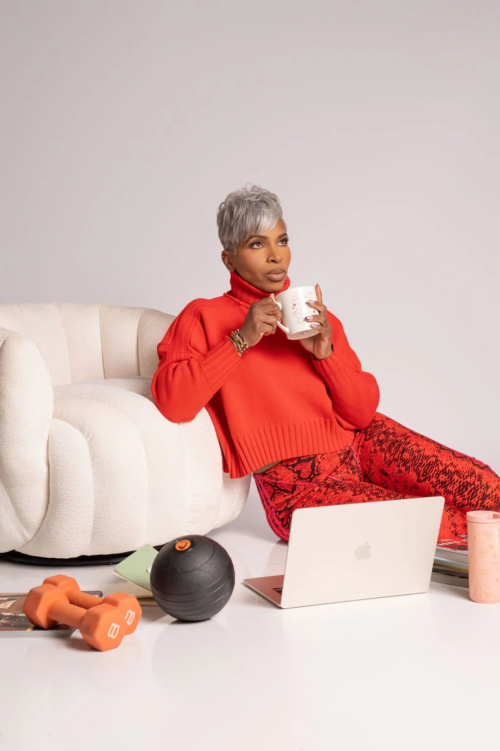 A woman with short gray hair sitting on the floor, holding a mug, surrounded by workout equipment, a laptop, and books, with a white couch in the background.