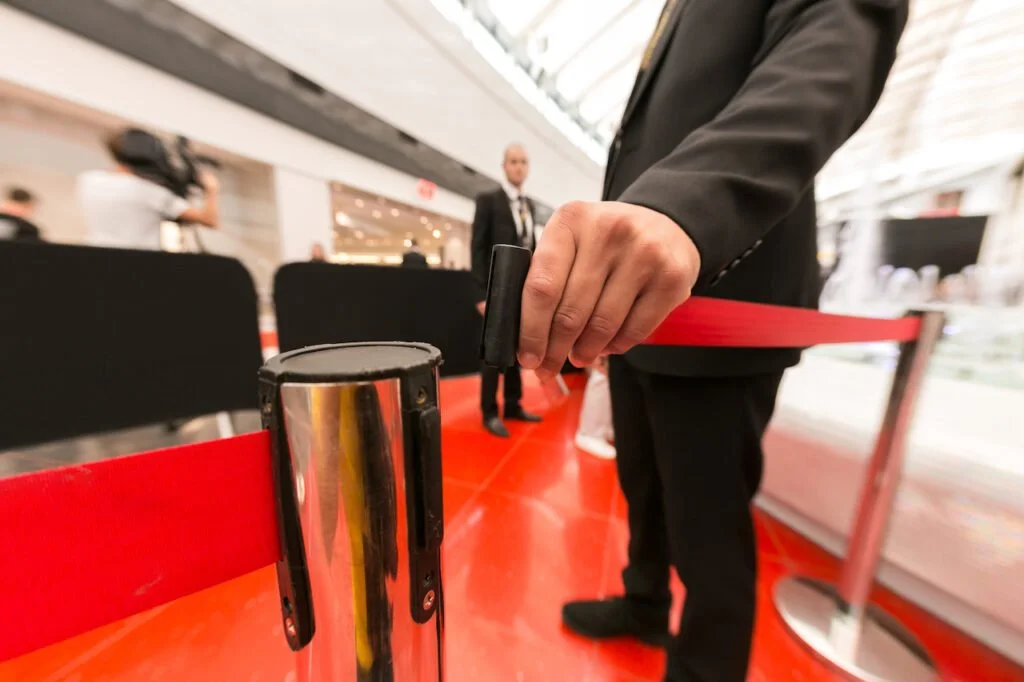 A person in a suit is pulling a red retractable barrier rope in what appears to be an airport or convention center. The focus is on the barrier's post and the person's hand.