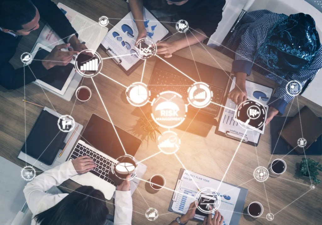 Business meeting at a wooden table with laptops, tablets, documents, and coffee cups, overlaid with interconnected icons related to risk management and data analysis.