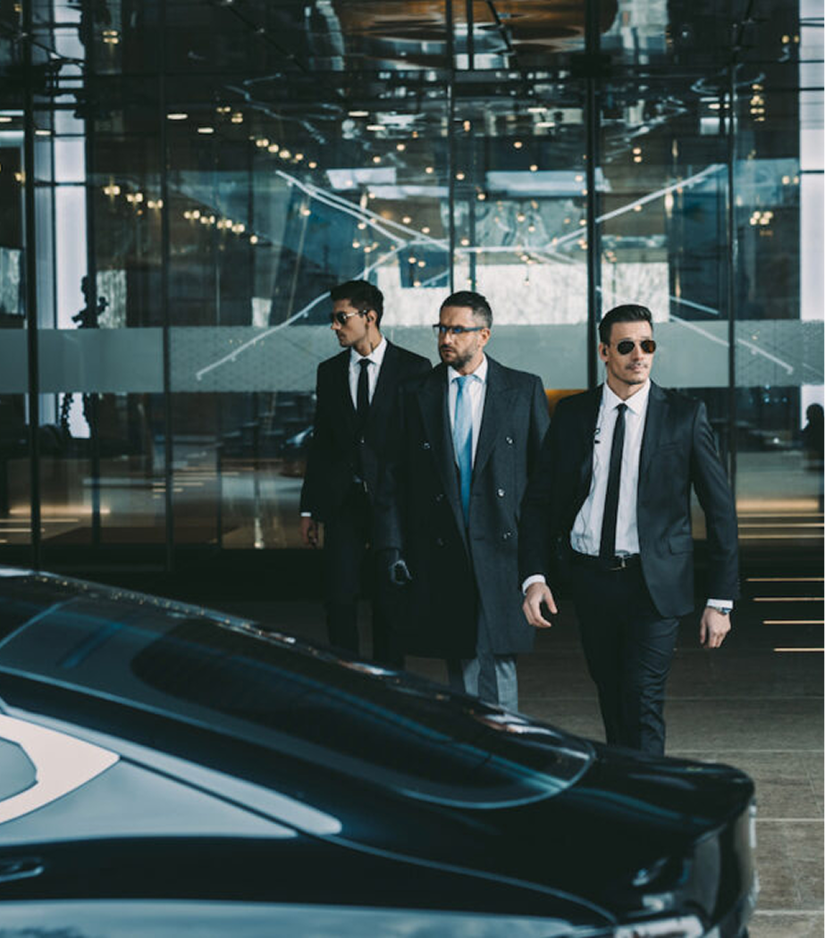 Three men dressed in suits walking outside a modern glass building, possibly a hotel or office, with a luxury car in the foreground.