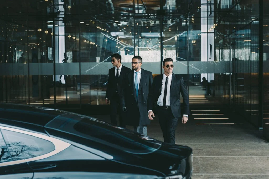 Three men in suits walking out of a modern building, with a black car in the foreground.