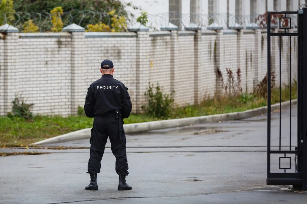 A security guard in all black uniform standing outside near a gated entrance, with a brick wall and some plants in the background.