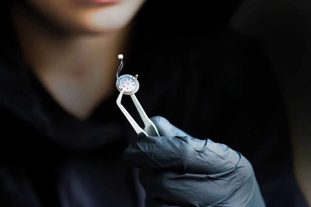 Close-up of a person wearing black gloves holding a small, metallic, circular dental or medical instrument with a handle and attached wires, against a dark background.