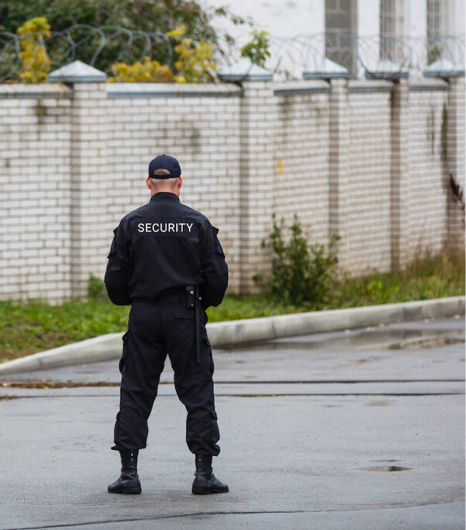 Security guard in black uniform standing on a wet street, facing away from the camera, near a brick wall with a barbed wire fence on top.