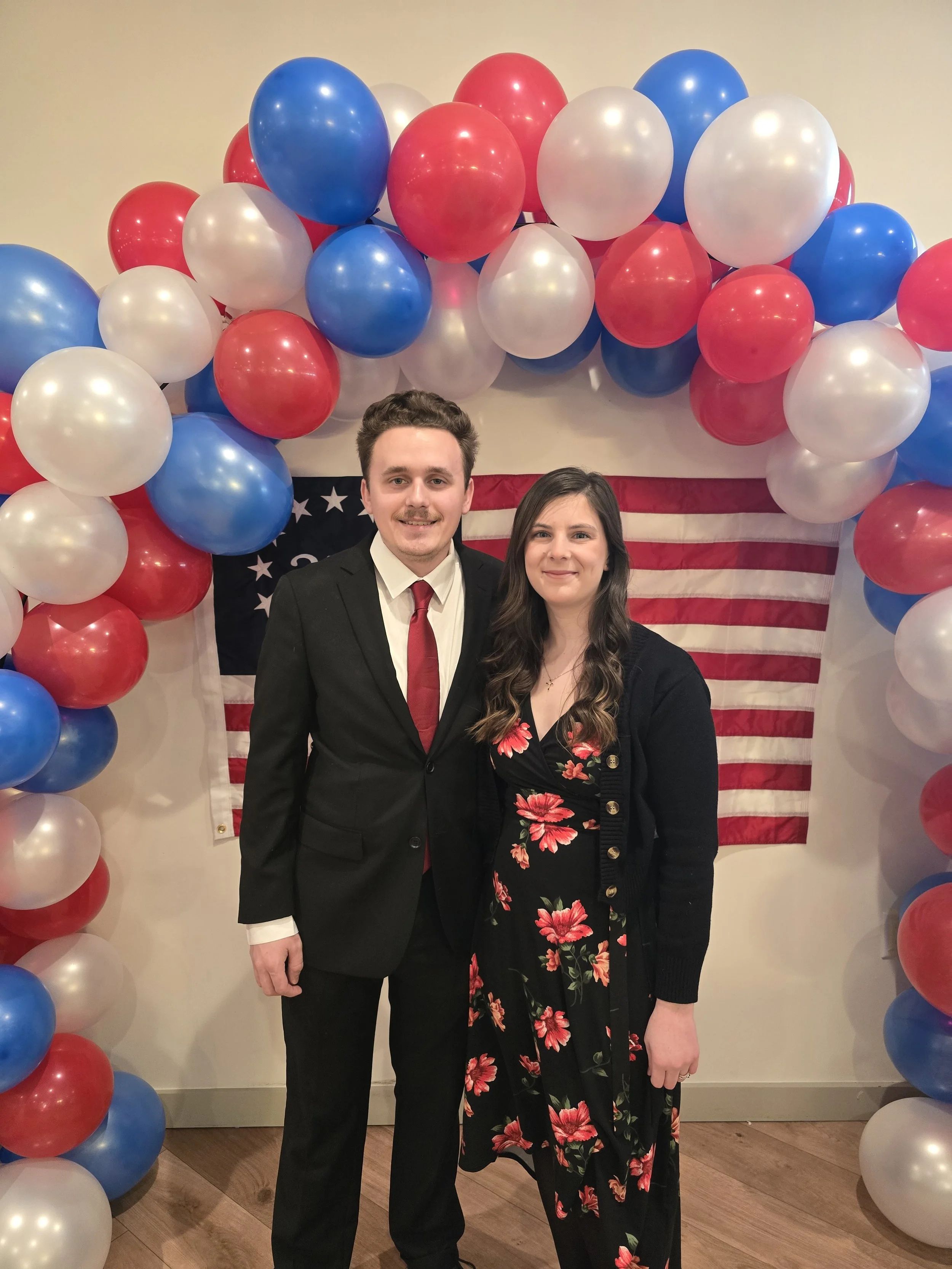 A young man and woman standing together at a patriotic event with red, white, and blue balloons and an American flag backdrop behind them.