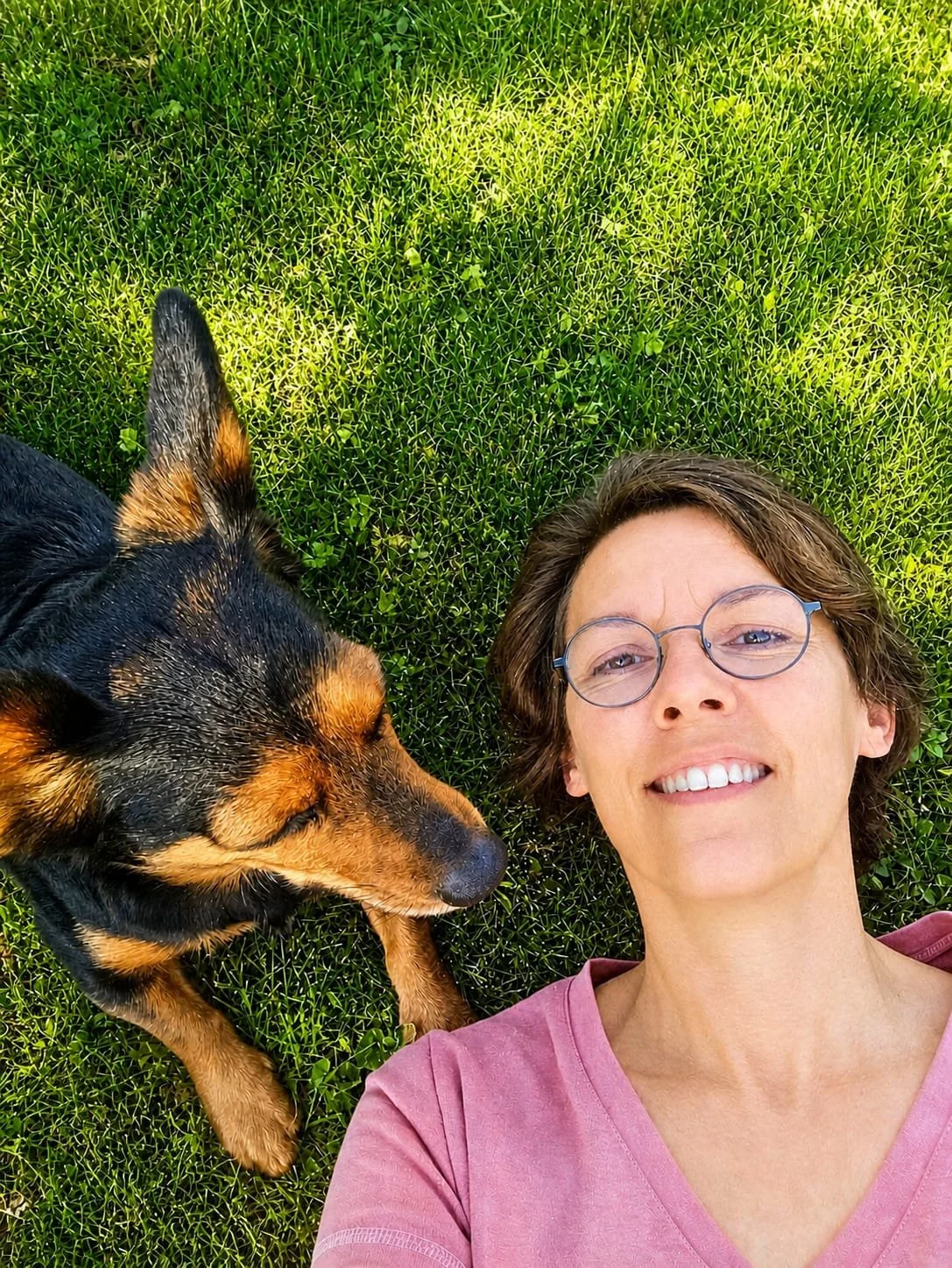 Une femme avec des lunettes souriant allongée sur l'herbe verte avec un chien noir et marron.