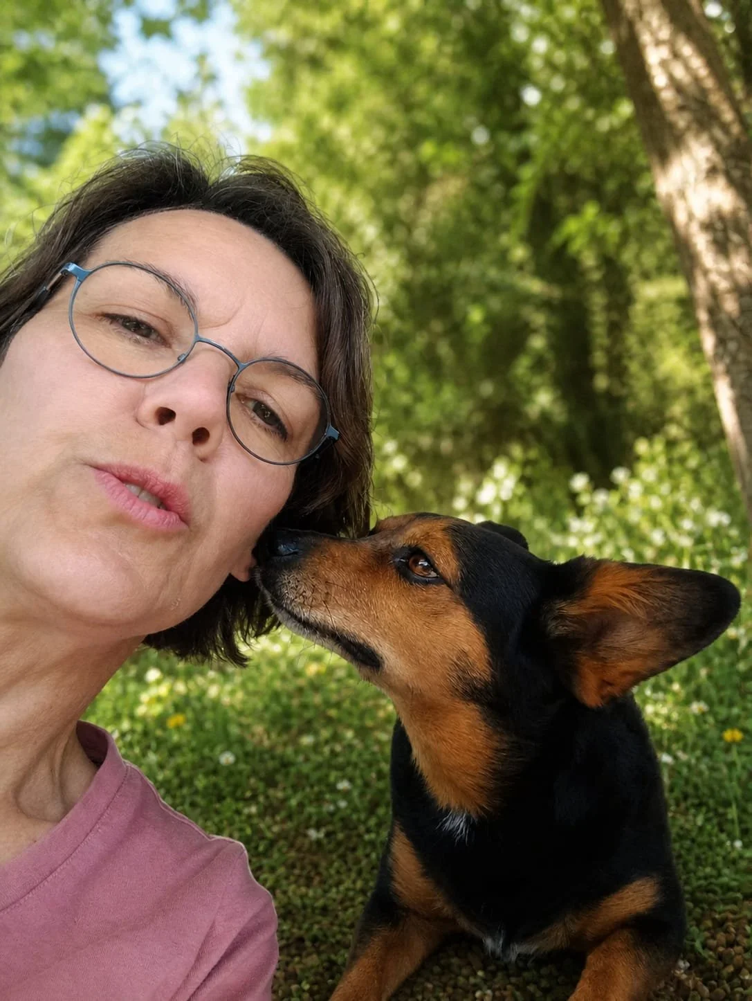 Une femme portant des lunettes et un t-shirt rose nez à nez avec un chien noir et marron dans un parc avec des arbres en arrière-plan.