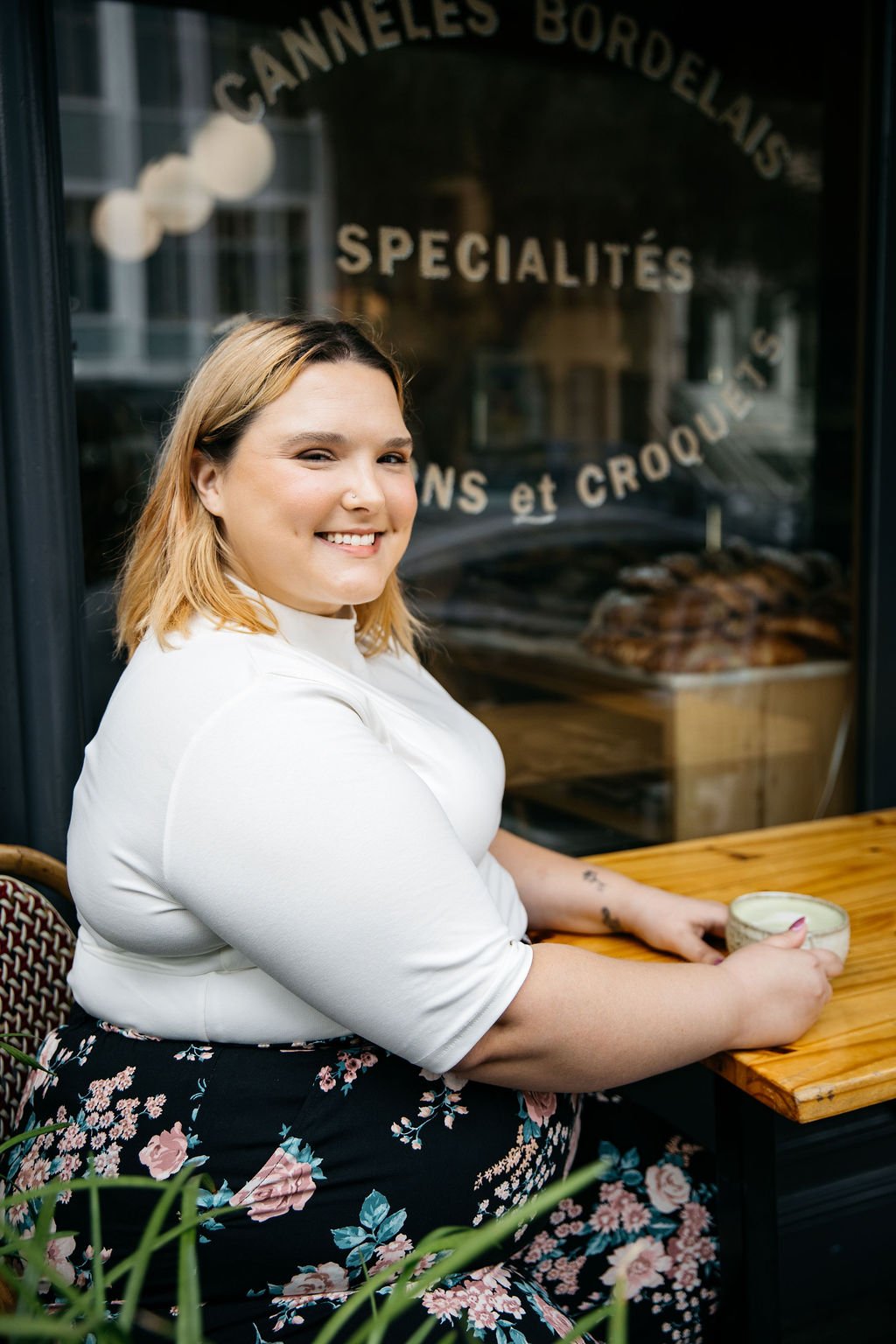 A woman sitting at a wooden table outside a bakery, smiling and holding a cup of coffee or tea, with a bakery window behind her showing baked goods and French writing.