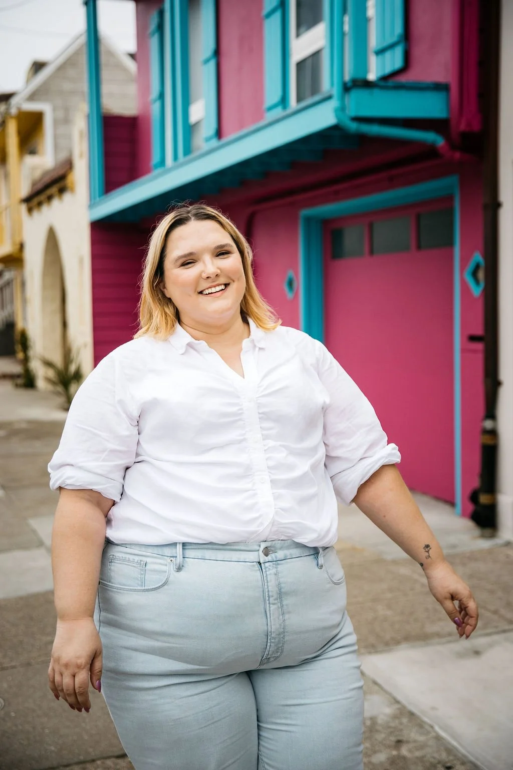 A smiling woman with light brown hair, wearing a white shirt and light blue jeans, standing on a colorful street with pink and blue houses in the background.
