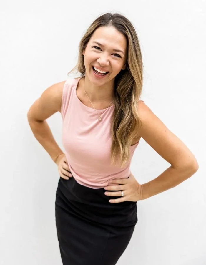 A woman in a pink sleeveless top and black skirt smiling with hands on her hips against a plain white background.