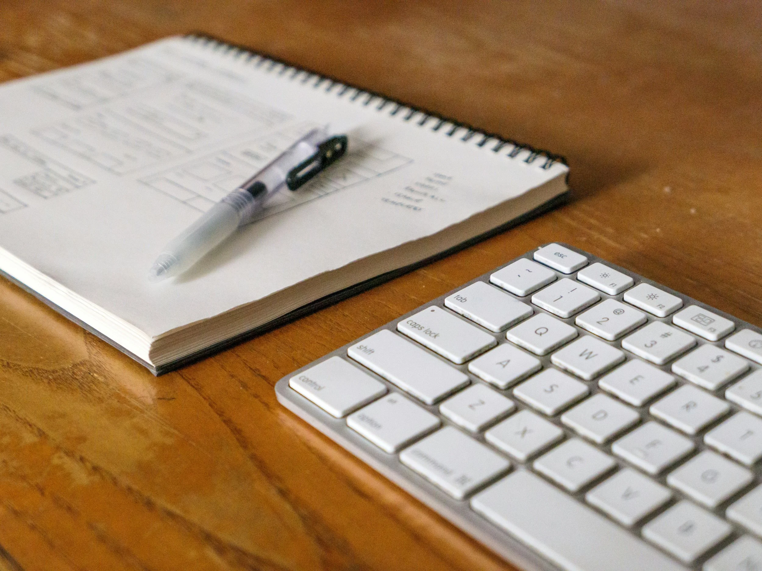 A spiral notebook with diagrams and notes, a black and clear pen, and part of a white computer keyboard on a wooden desk.