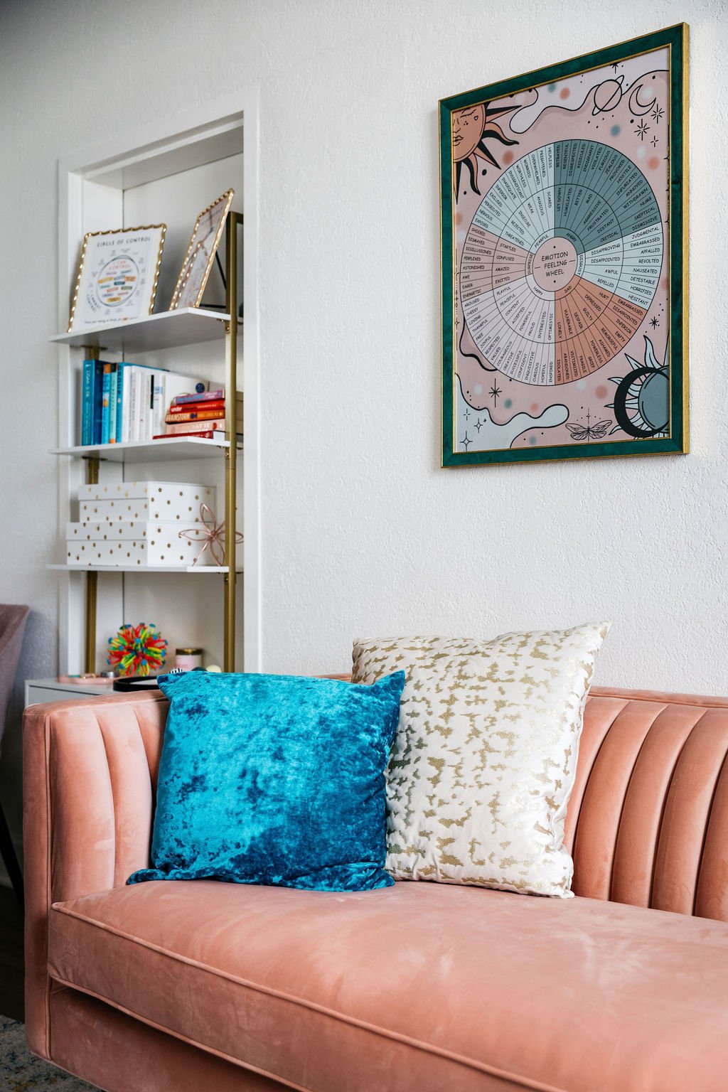 Living room with a peach velvet sofa, two decorative pillows in blue and cream, a white bookshelf with books and decorative items, and a framed poster of the emotion feeling wheel on the wall.