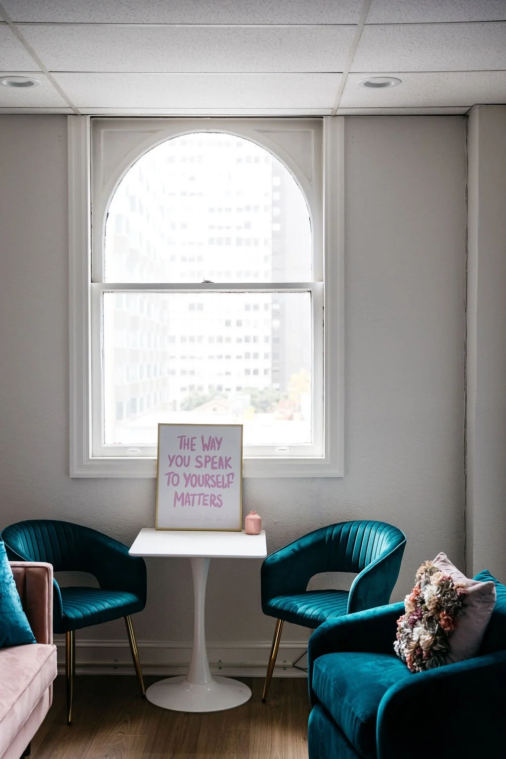 A cozy sitting area with two teal velvet chairs, a small white table, and a pink cushion with flowers. There's a framed quote on the table that reads, "The way you speak to yourself matters." Behind the table is a large arched window with a cityscape view.