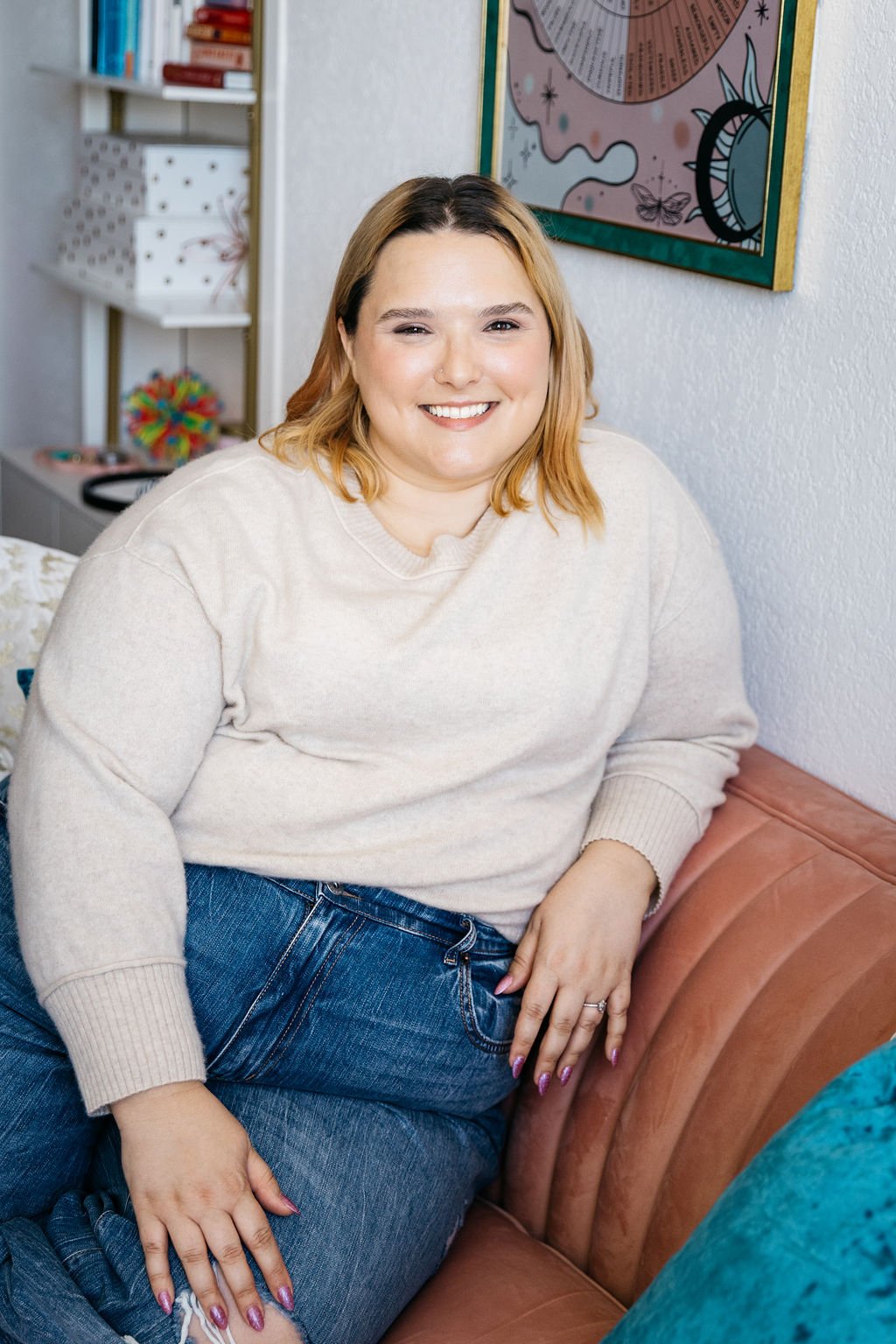 A smiling woman with shoulder-length blonde hair sitting on a pink sofa, wearing a cream sweater and blue jeans, in a room with a white wall, bookshelf, and colorful wall art.