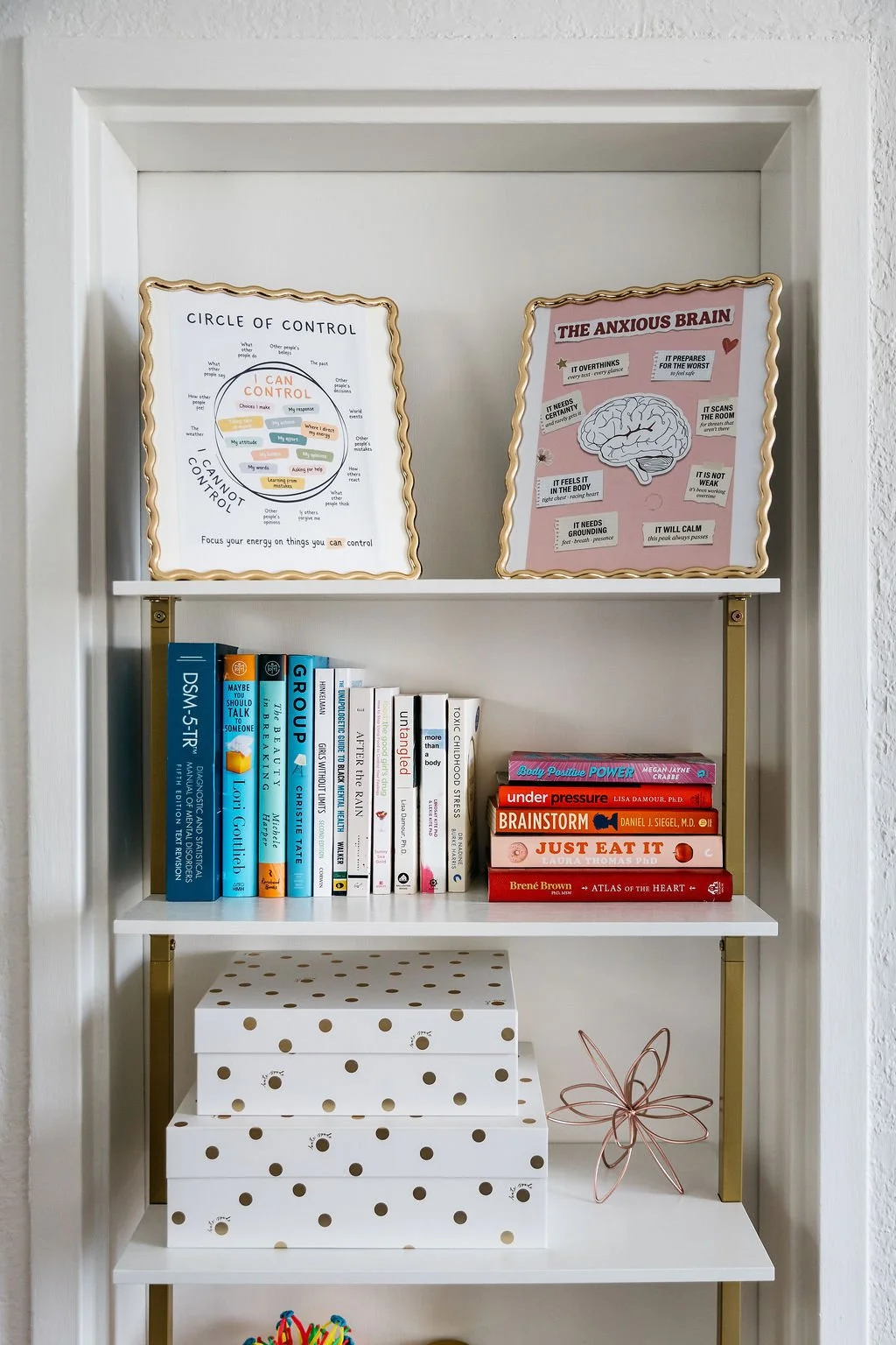 A white bookshelf with two framed posters at the top, one titled 'Circle of Control' and the other 'The Anxious Brain.' Below the posters, there are books of various colors and titles, some stacked horizontally and some vertically. There are two decorative white boxes with gold polka dots on the middle shelf and a wire flower-shaped decor piece beside them.