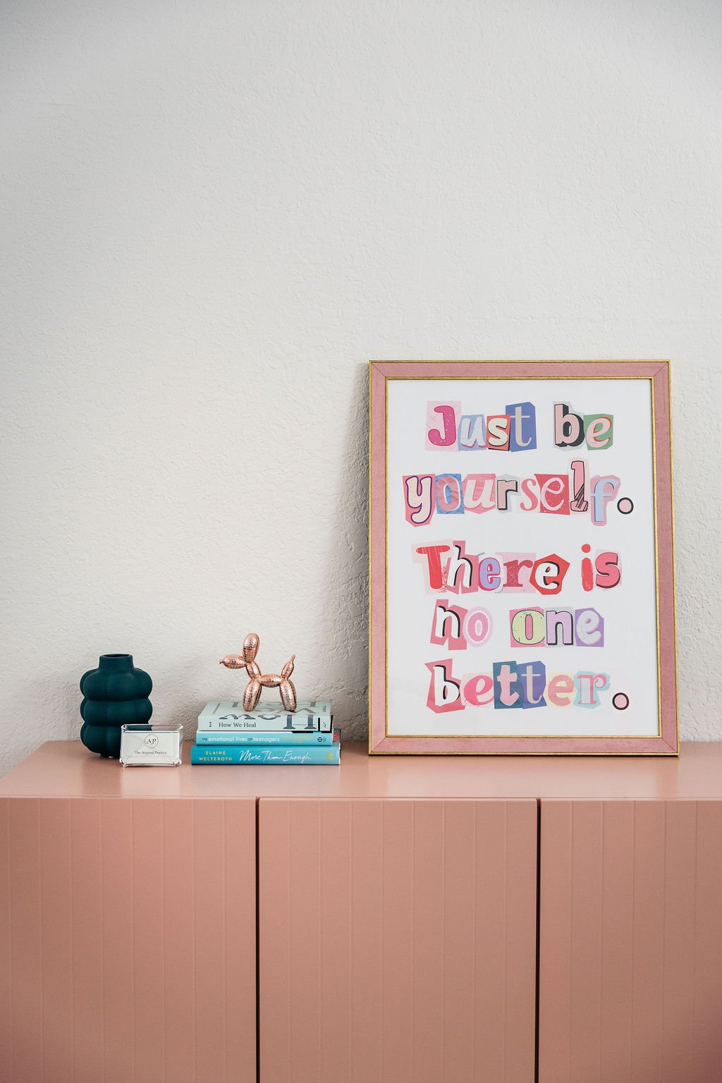 Decorative pink cabinet with a framed inspirational quote, a teal vase, a small pink balloon animal, and three stacked books on top.