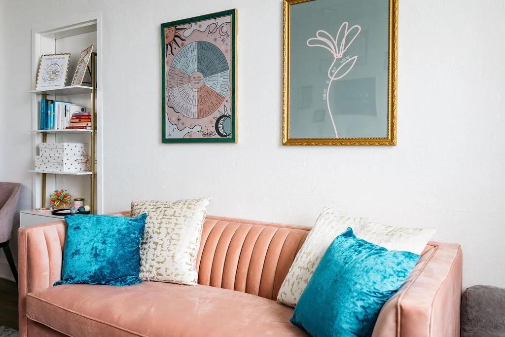 Living room with a pink velvet sofa, four pillows (two white and two turquoise), a white bookshelf with books and decorative items, and two framed artworks on the white wall above the sofa.