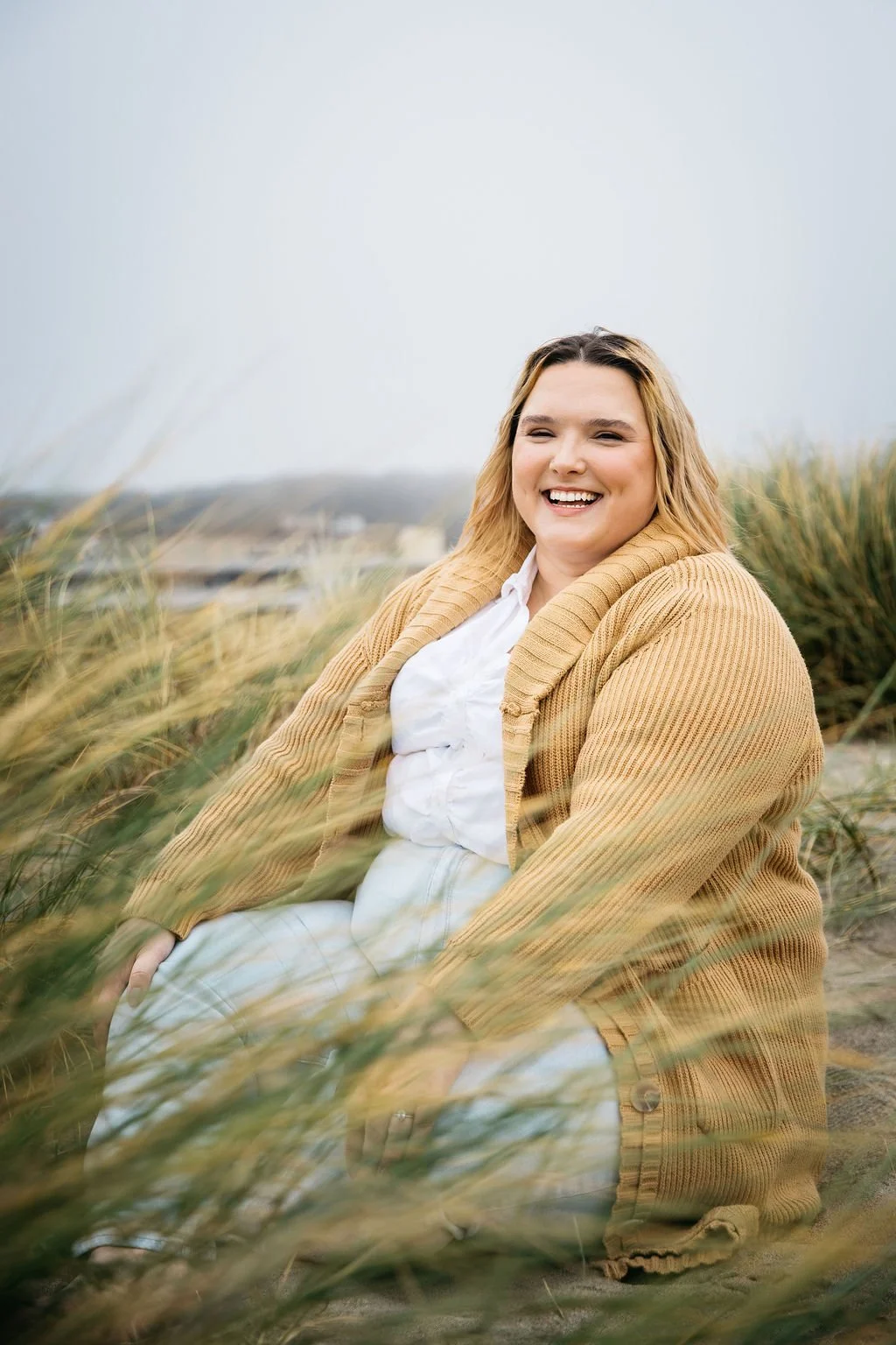 A woman sitting outdoors among tall grass, smiling and wearing a beige cardigan and white outfit, with a cloudy sky in the background.