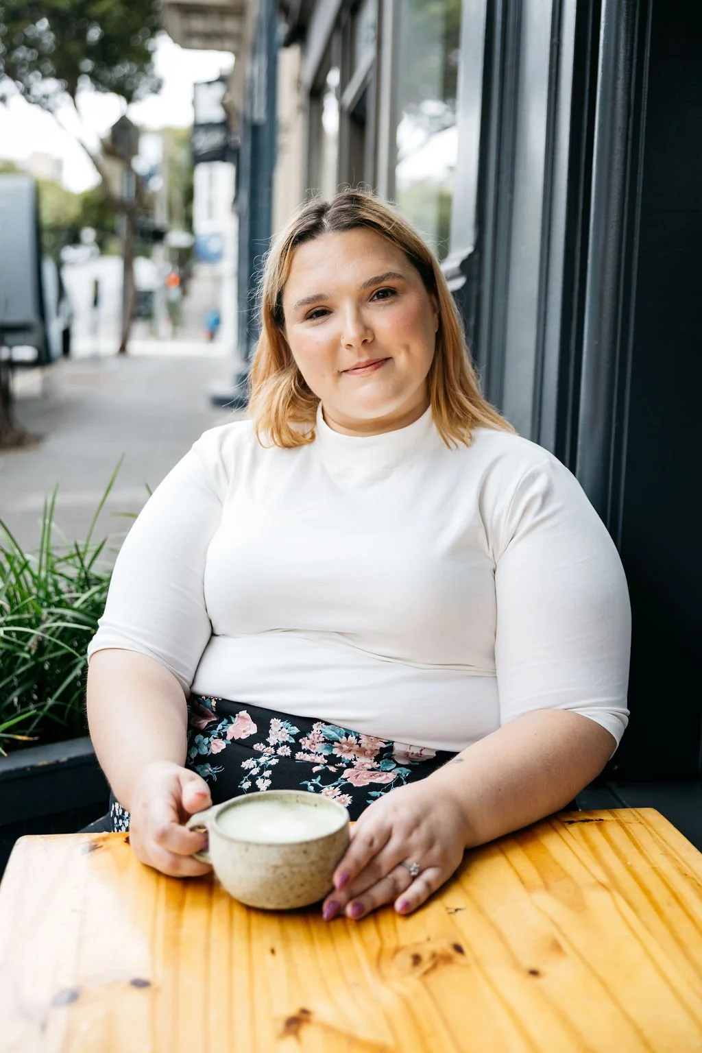 A woman with shoulder-length light brown hair, wearing a white top, sitting at a wooden table outside a cafe, holding a ceramic cup of coffee with a foam top.