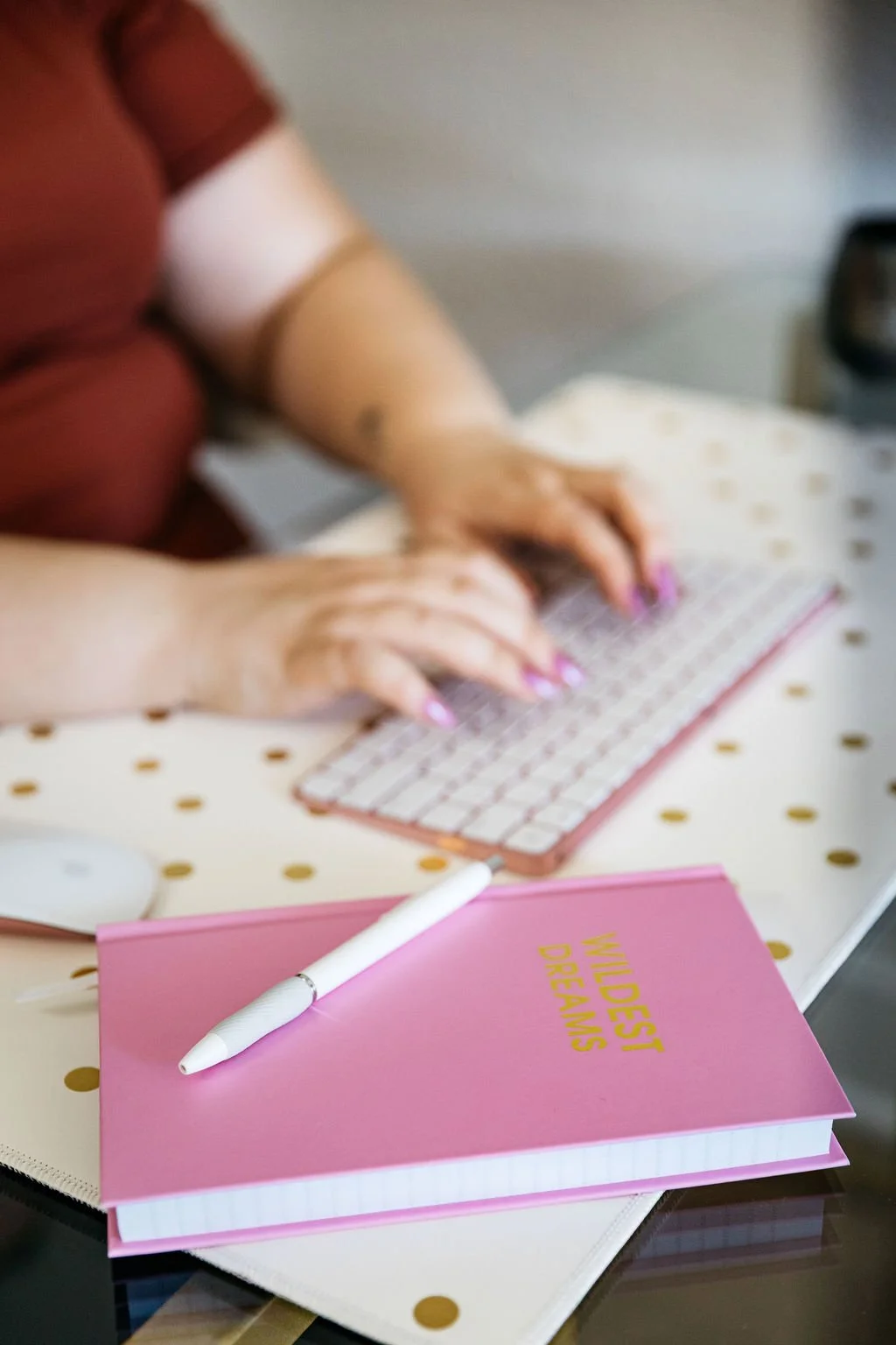 A person typing on a small, pink keyboard with a pink notepad labeled 'WILDEST DREAMS' and a white pen on top, sitting at a dotted table with a blurred background.