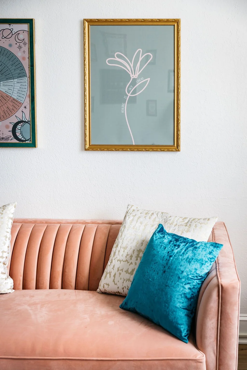 A pastel pink velvet sofa with three decorative pillows in white and bright blue, against a white textured wall with two framed artworks, one featuring a flower drawing with the phrase "HEALING IS NOT LINEAR".