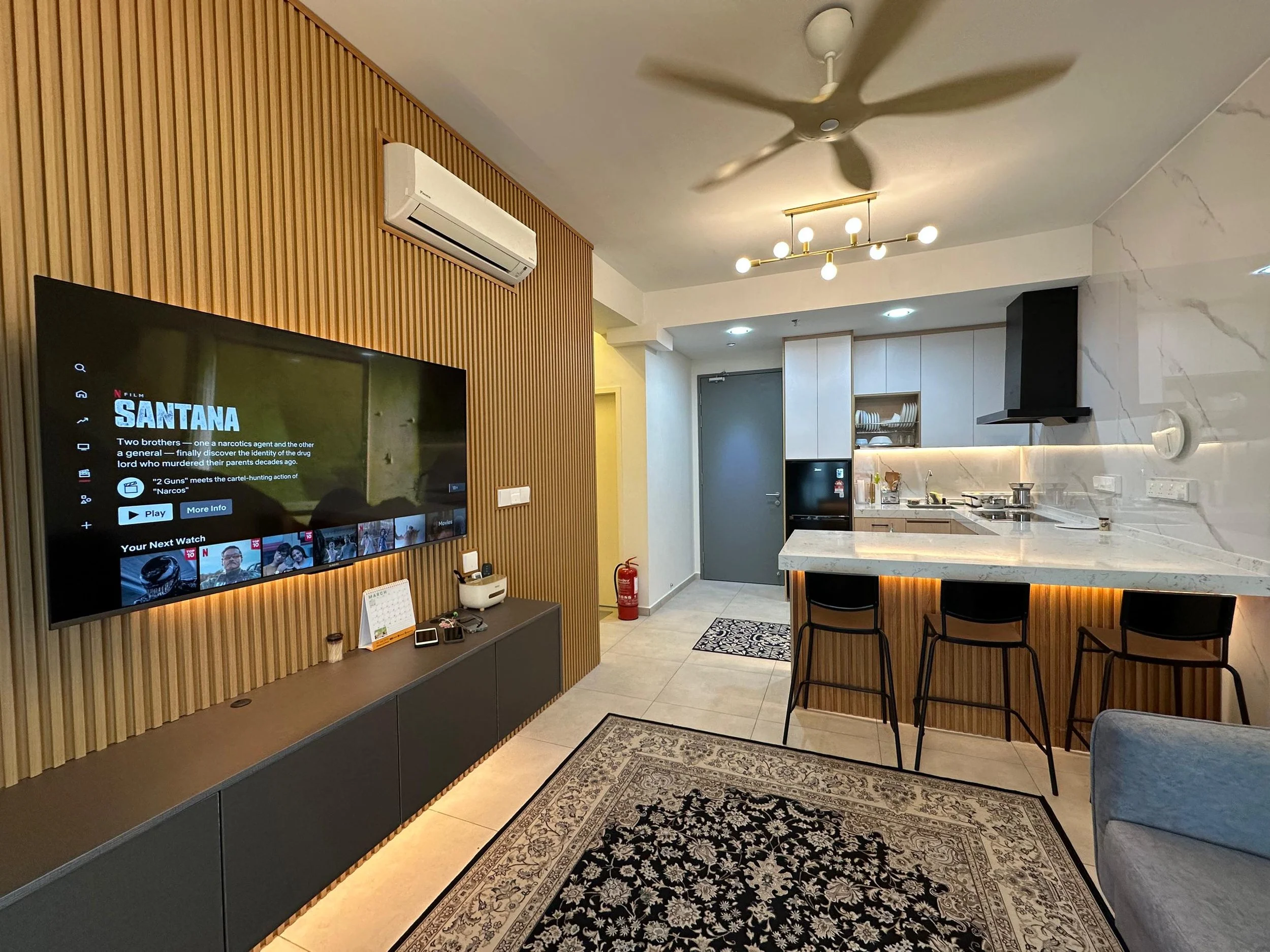 Modern living room with a mounted flat-screen TV on a wooden slat wall, a white air conditioner, a gold and globe chandelier, a kitchen with white cabinetry, a black range hood, and a marble island with chairs, a patterned rug, and a door in the back