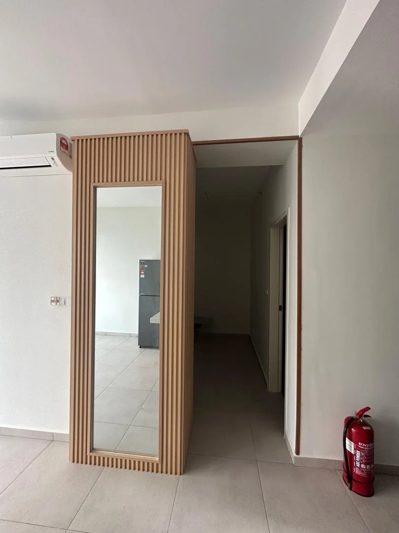 Interior room with a decorative wooden panel with a mirror, a white wall-mounted air conditioner, a fire extinguisher on the floor, and a doorway leading to a kitchen area with a silver refrigerator.