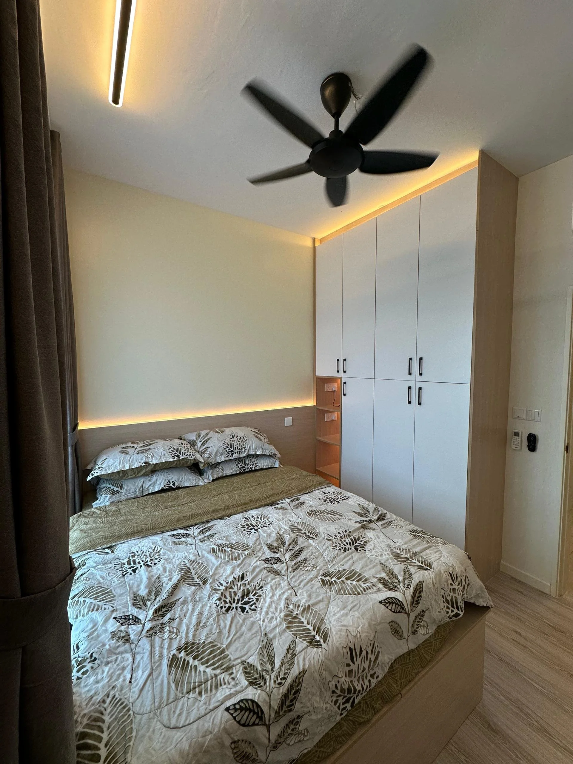 A bedroom with a bed, pillows, and a bedspread with leaf patterns. There is a built-in wardrobe with black handles and shelves, and a ceiling fan with dark blades. The room has soft lighting along the ceiling edges and appears modern and minimalistic