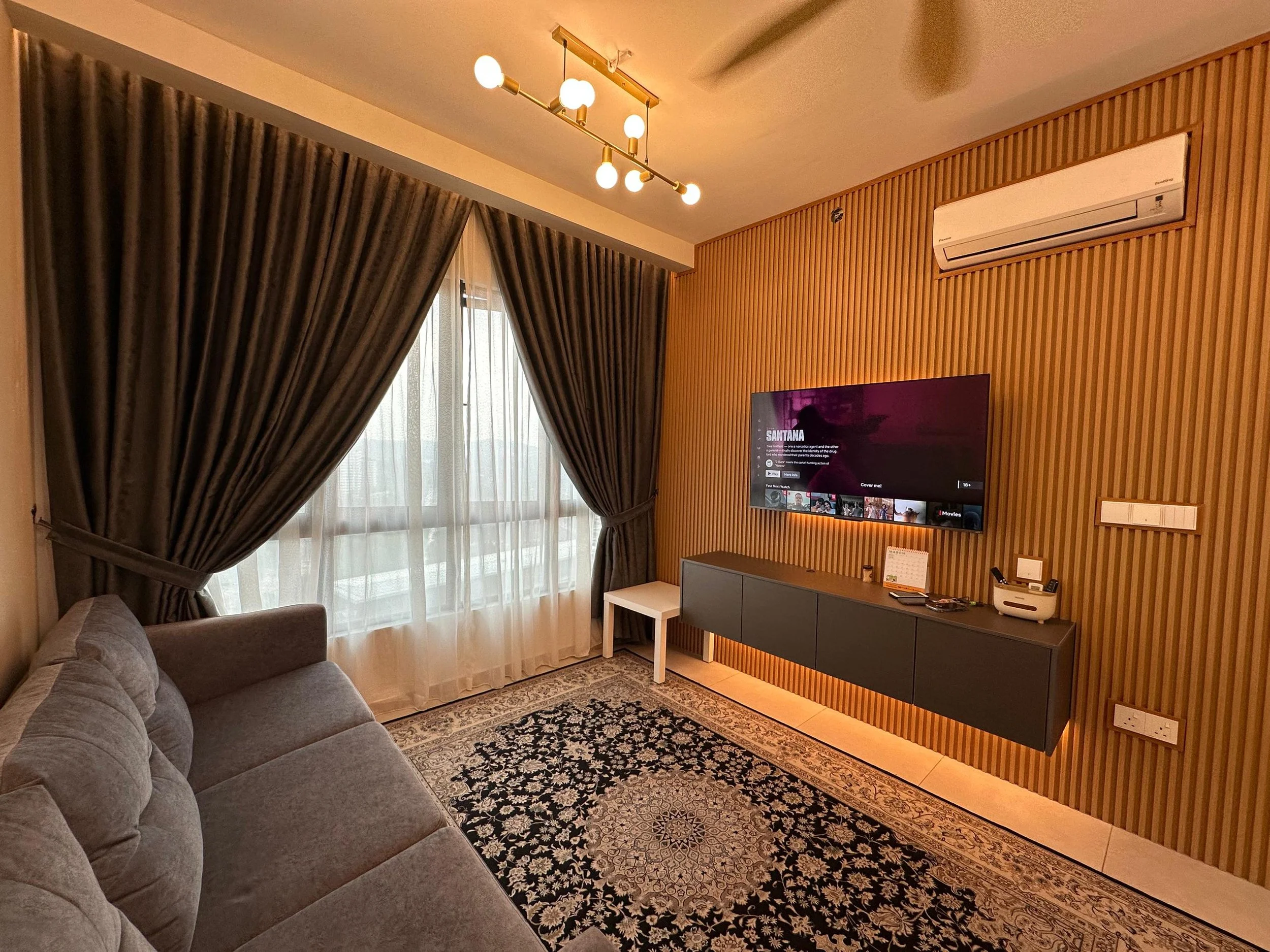 A living room with a gray sofa, a window with heavy brown curtains and sheer white curtains, a patterned rug, a wall-mounted television, and a light wooden wall panel with an air conditioning unit.