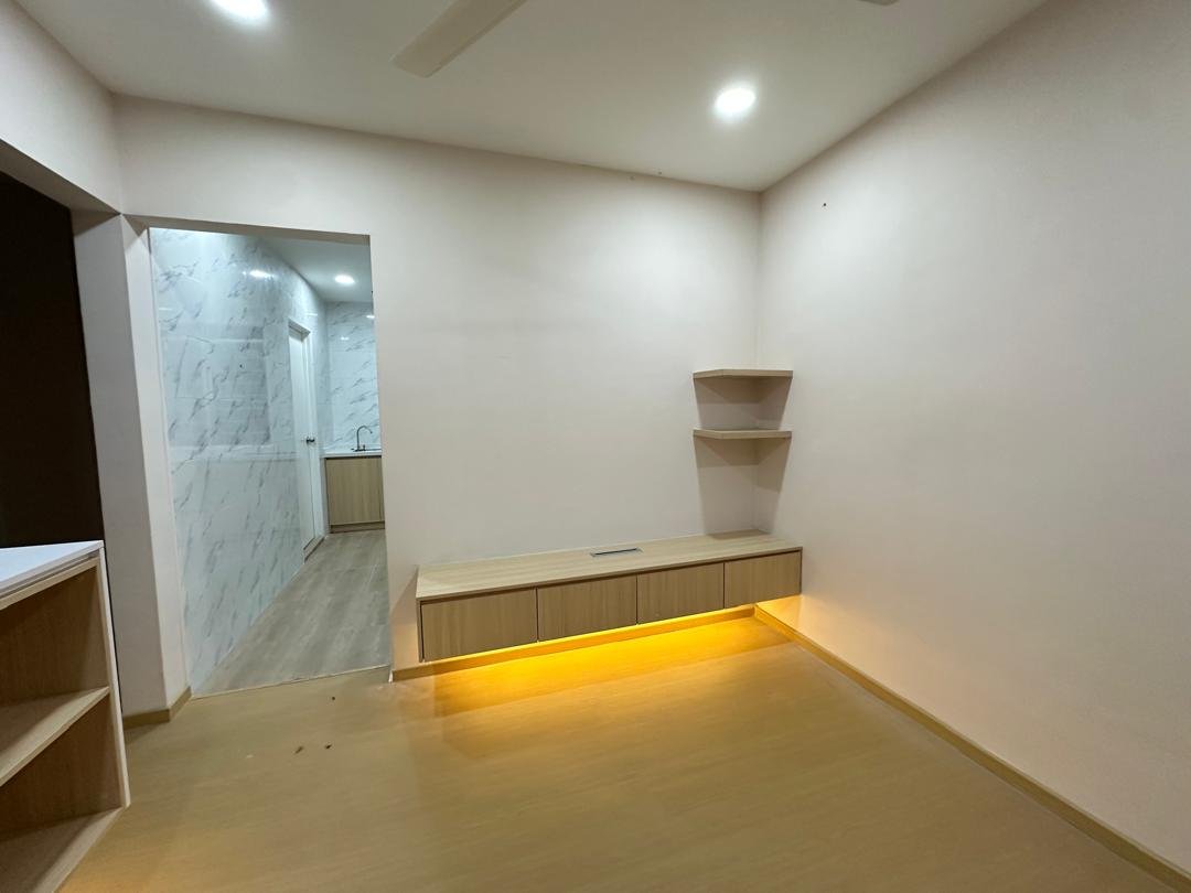 Empty living room with built-in wooden shelves and a low cabinet with warm yellow lighting, opening to a hallway with marble walls and a small kitchen area.