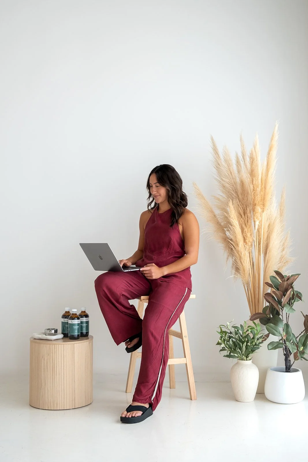 Woman in sleeveless maroon outfit sitting on a wooden stool working on a laptop in a minimalist room with potted plants and bottles on a round wooden table.