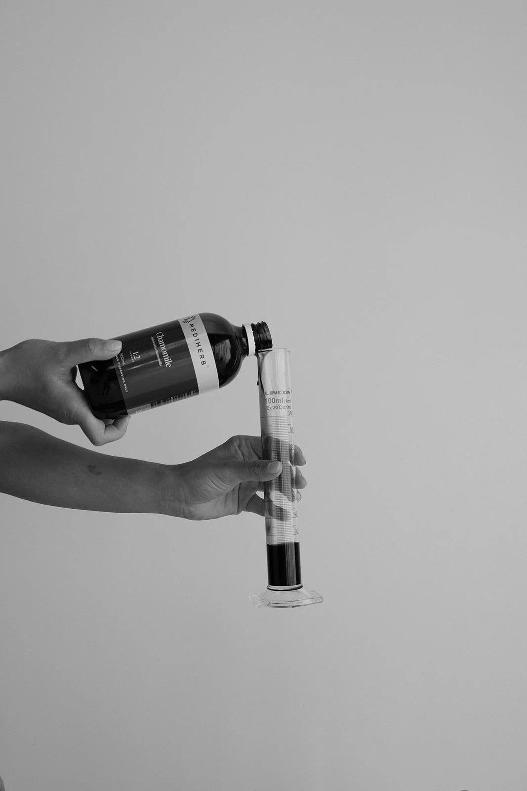 Black and white photo of a person pouring herbal liquid from a bottle into a graduated cylinder