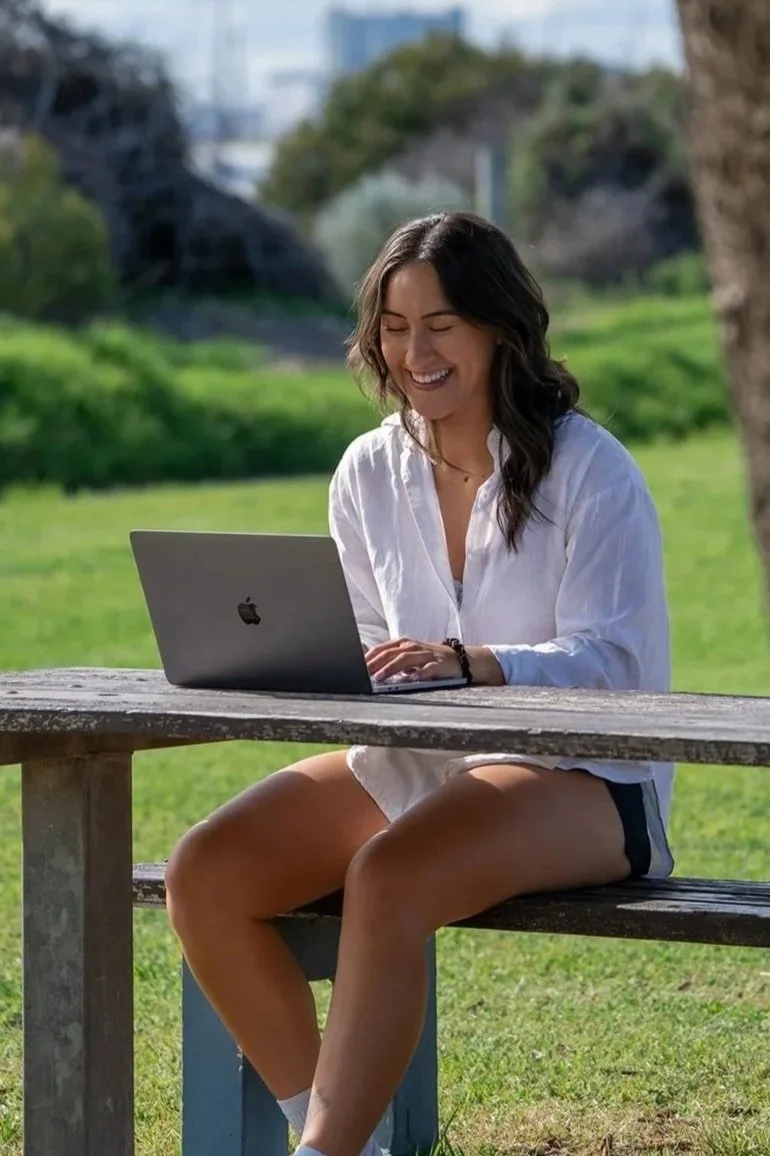 Young woman sitting at a picnic table outdoors using a laptop, smiling, with green grass and trees in the background.