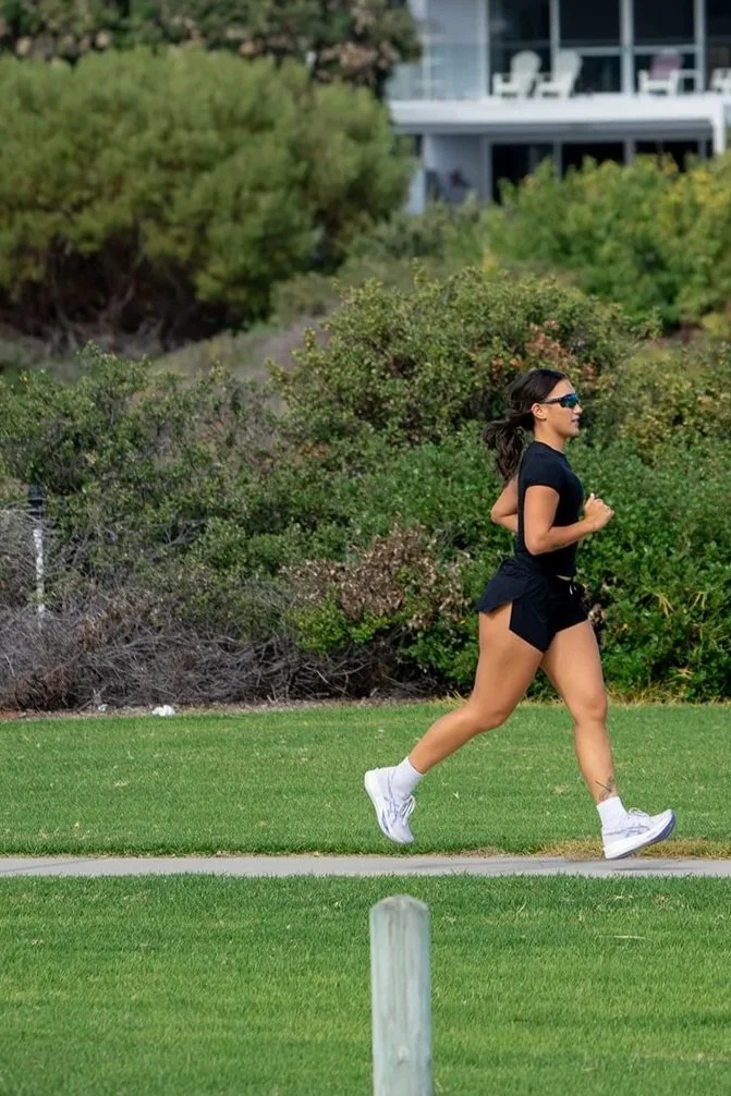 A woman running outdoors on a grassy path, wearing black athletic clothes, white sneakers, and sunglasses, with greenery and a building in the background.