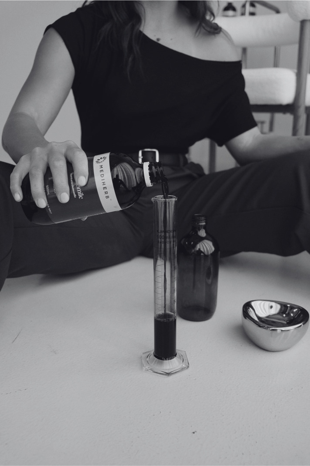 A person pours a herbal liquid from a labeled bottle into a graduated cylinder. The person is sitting cross-legged on the floor, wearing a black off-the-shoulder top. There is a small metal bowl and another bottle on the floor.