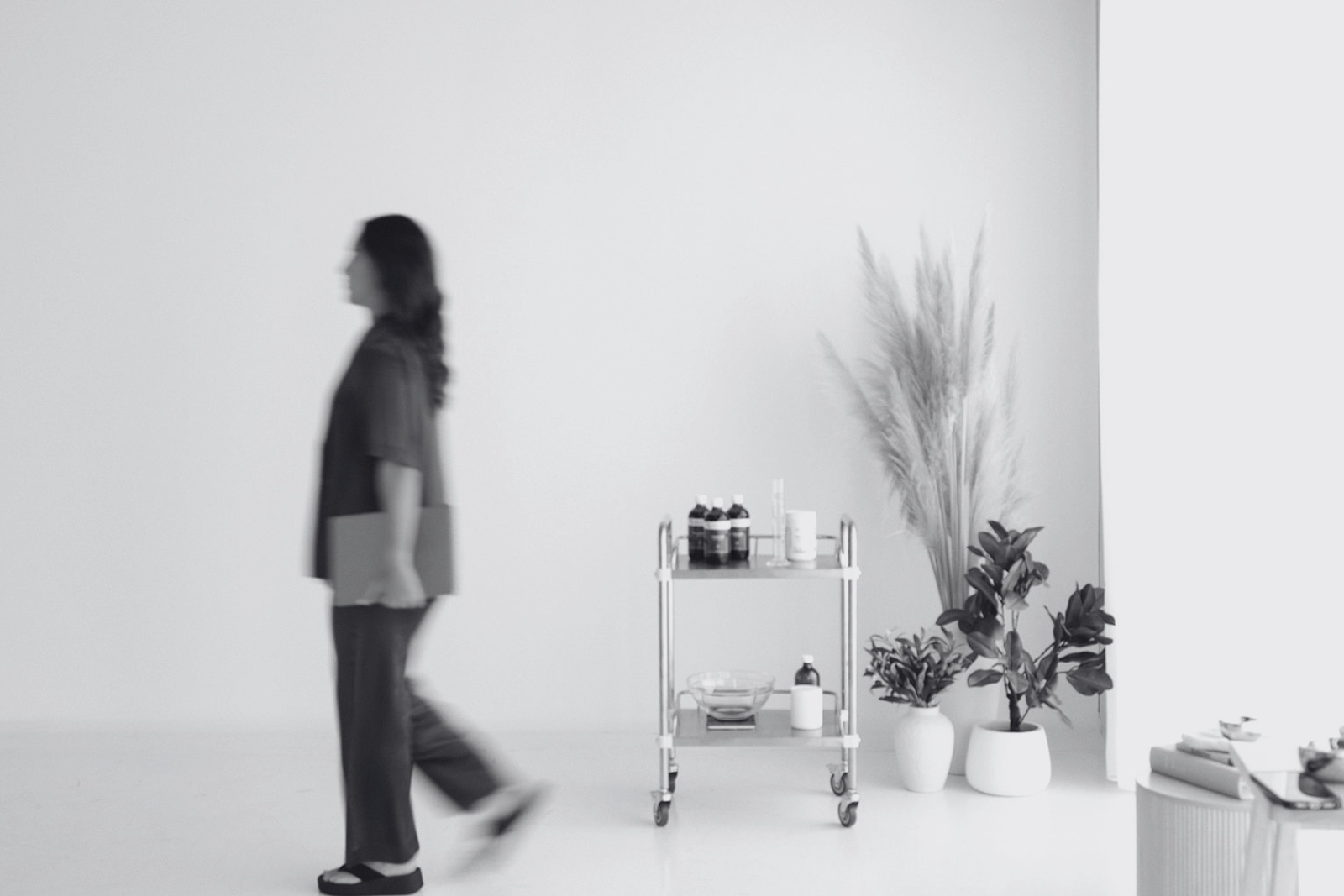 A woman walking past a minimalist white space with a small cart holding bottles and jars, and potted plants near a window.