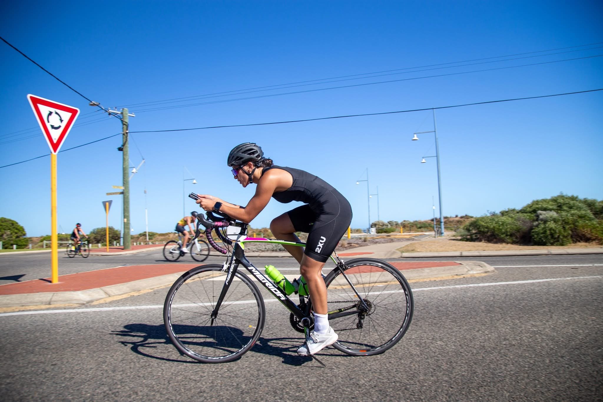 Cyclist wearing a black helmet and sleeveless black sportswear riding a black and green bike on a paved road, with other cyclists in the background, clear blue sky, and a triangular traffic sign indicating a roundabout.