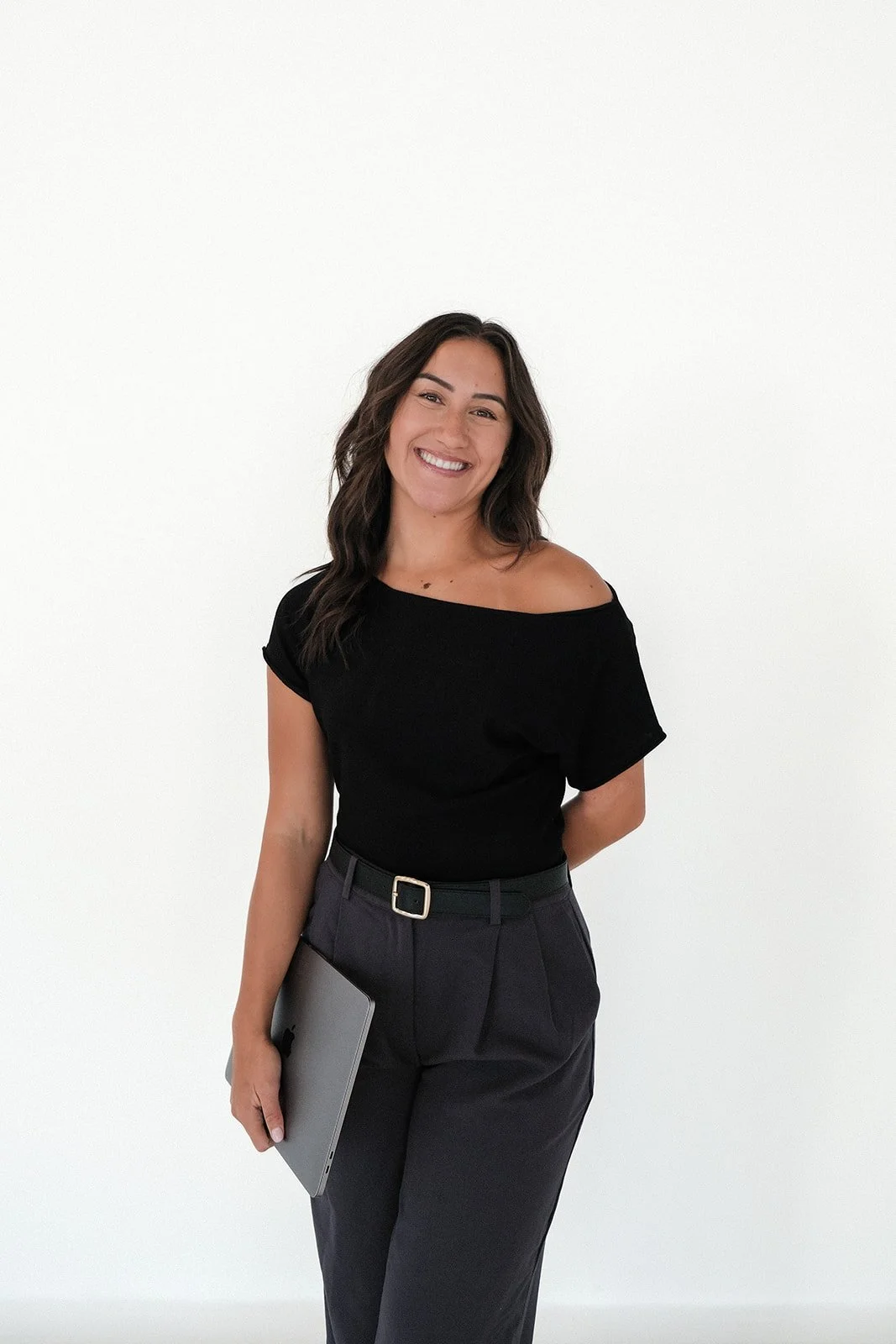 A woman with shoulder-length dark hair, smiling, wearing a black off-the-shoulder top and dark gray pants, holding a laptop, standing against a white background.