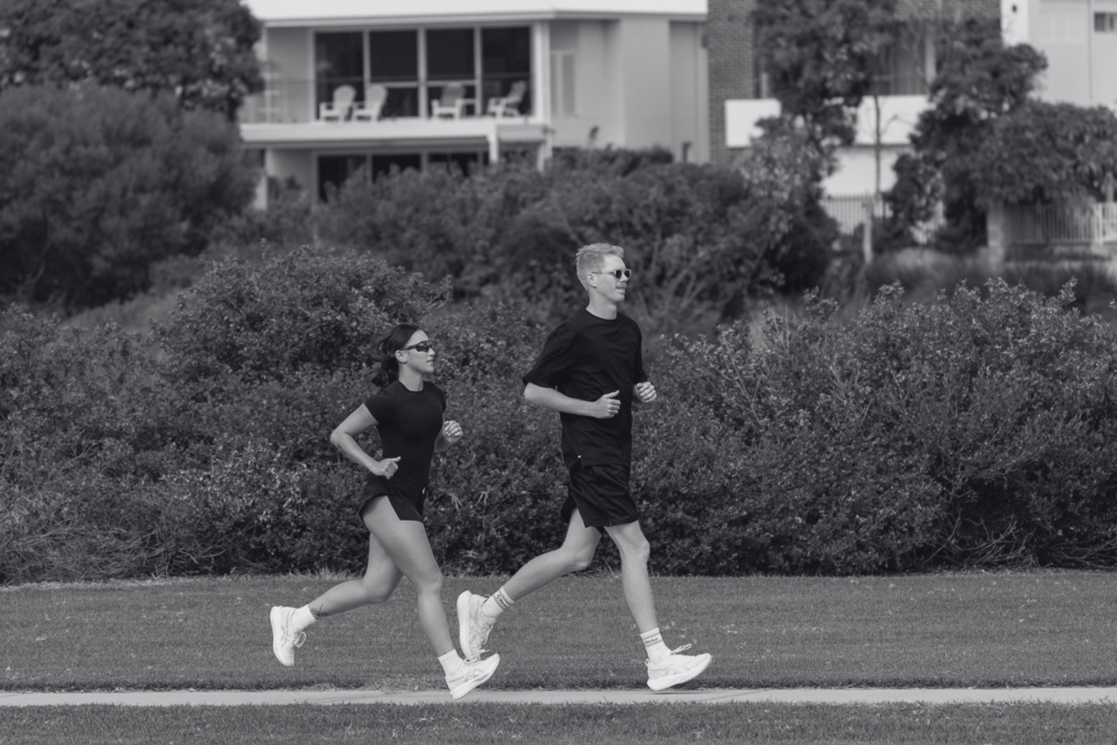 Two people, a man and a woman, jogging on a grassy area with shrubs and trees in the background. Both are wearing athletic clothing and sunglasses.