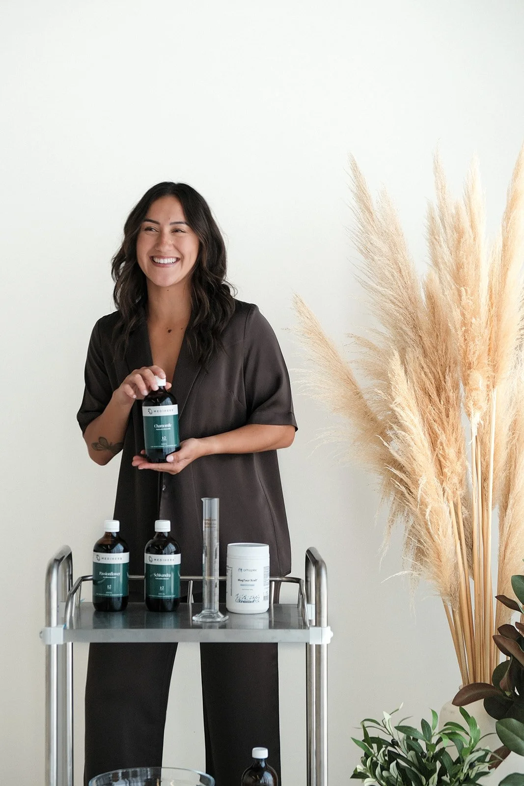 A woman with dark wavy hair holding a bottle of herbal medicine while standing next to a cart with similar bottles and a white container. She is smiling and standing indoors with a white wall and tall beige pampas grass in the background.