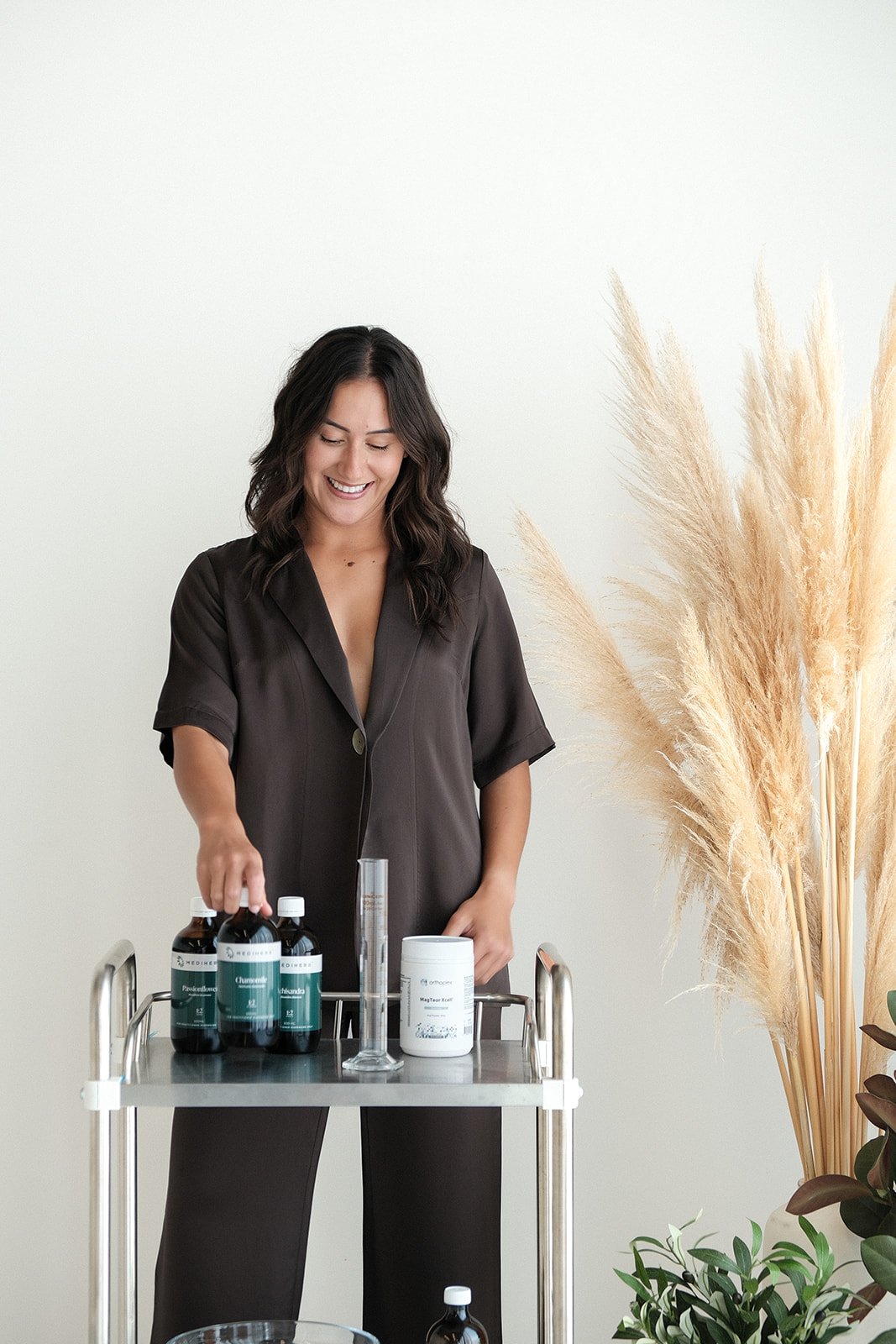A woman with dark hair smiling and standing behind a metal trolley with herbal medicine in a bottle, a graduated cylinder, and a container, next to tall pampas grass and green plants.
